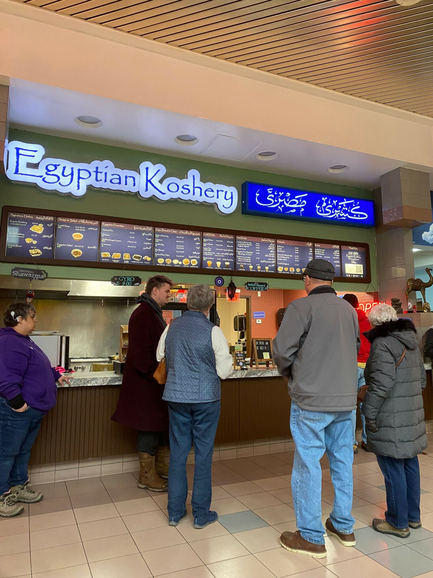 People standing in line at a counter inside a store or cafe, with a digital menu display above the counter.