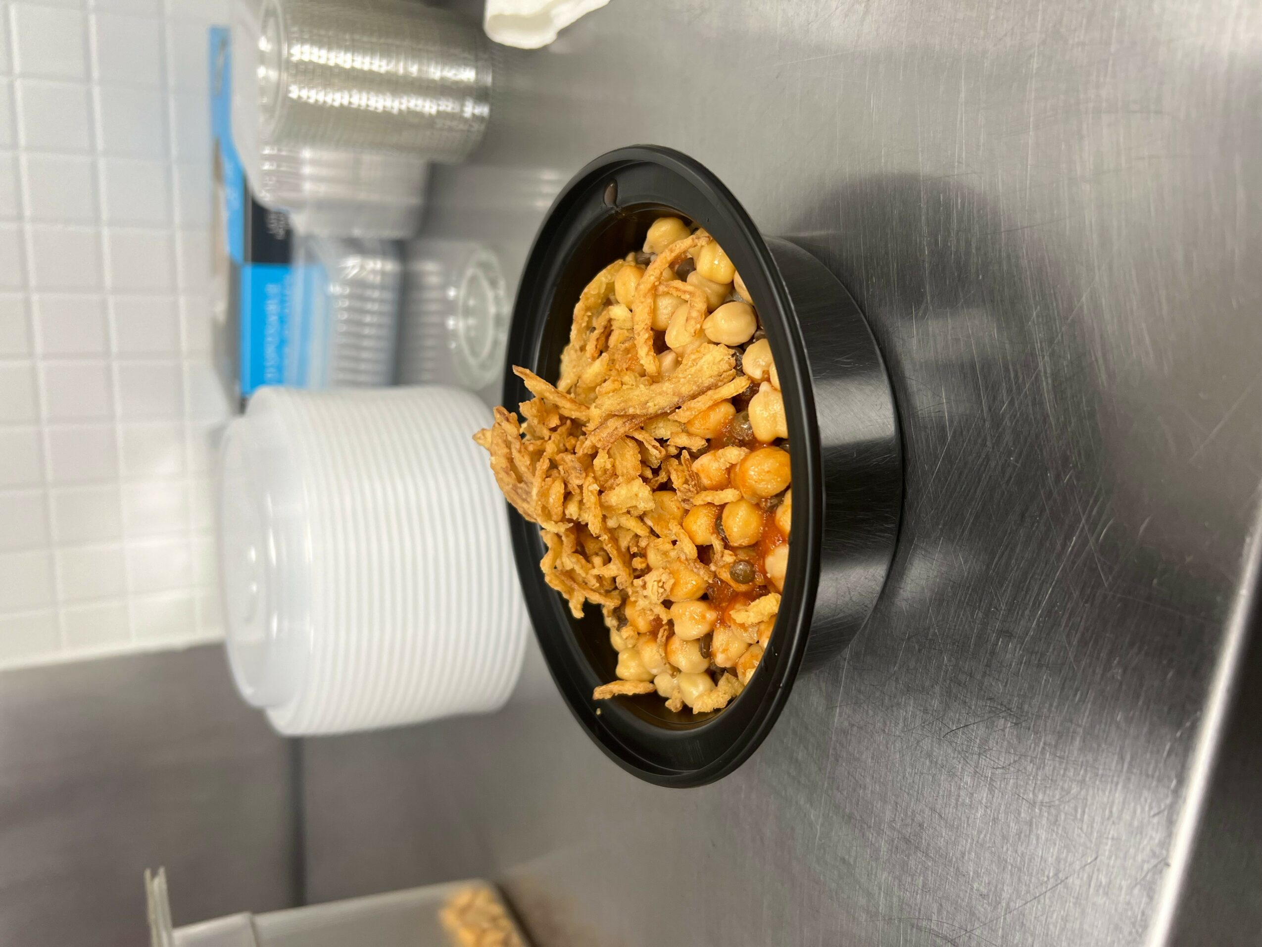 Bowl of fried chickpeas and crispy snack on a stainless steel surface, with stacked white bowls and glasses in background.