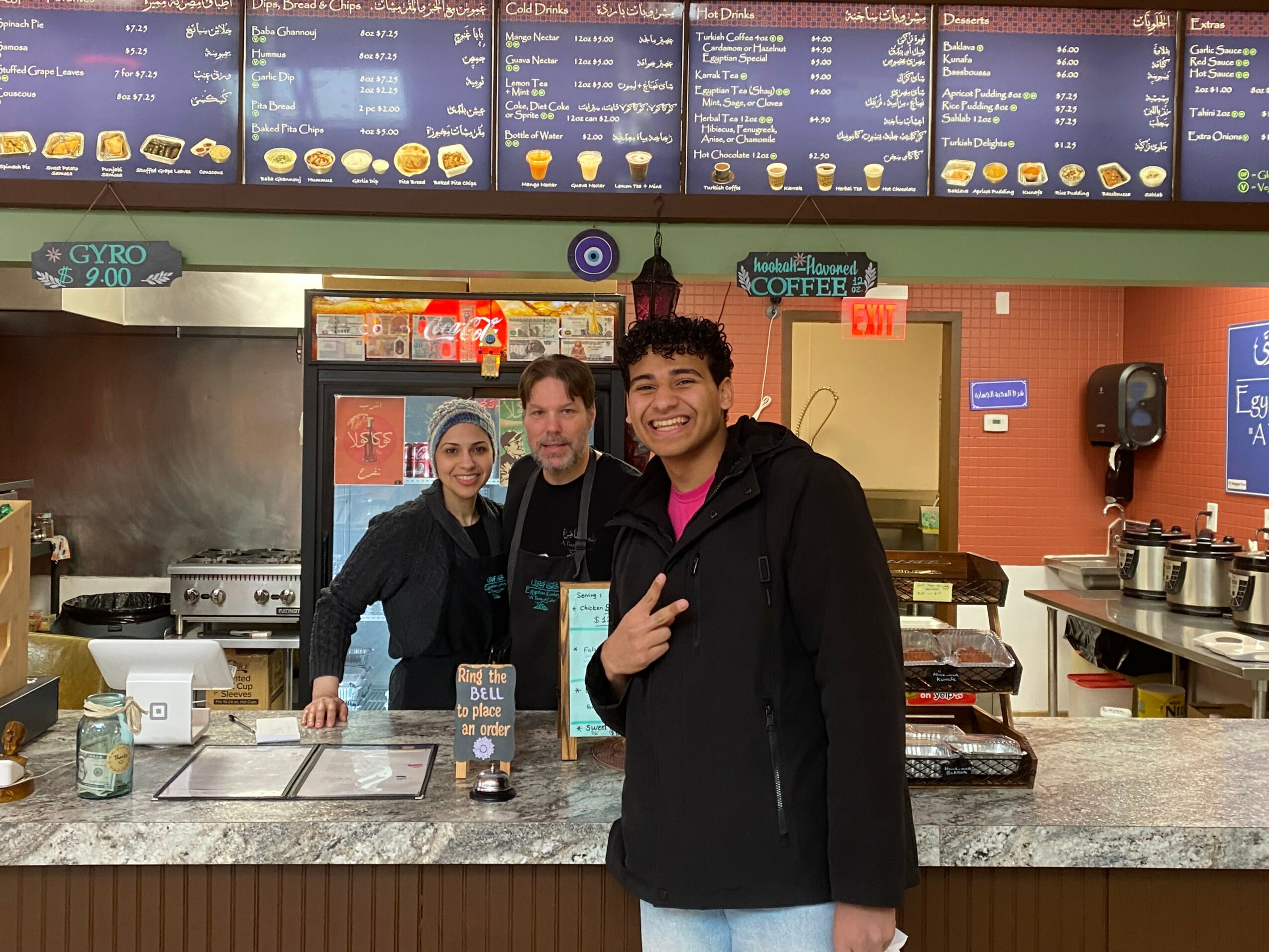Three people behind a counter in a restaurant, smiling, with menu boards above and kitchen equipment behind them.