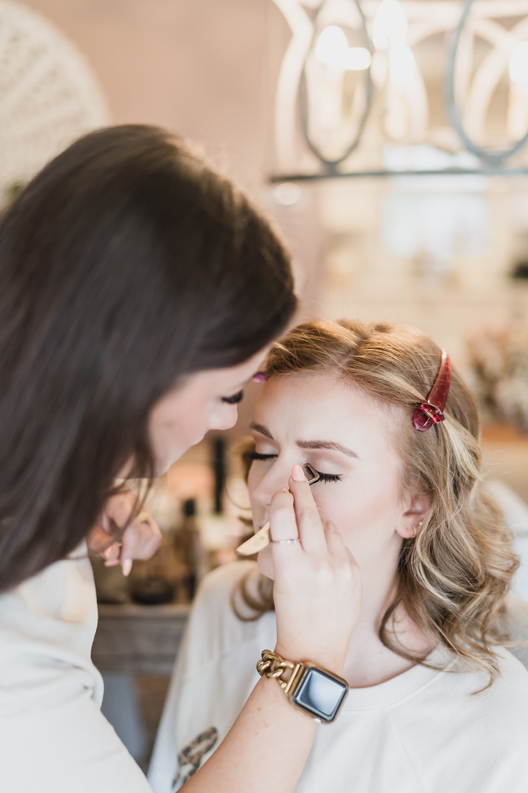 Woman applying makeup to a young girl with a hair clip, in a well-lit room.