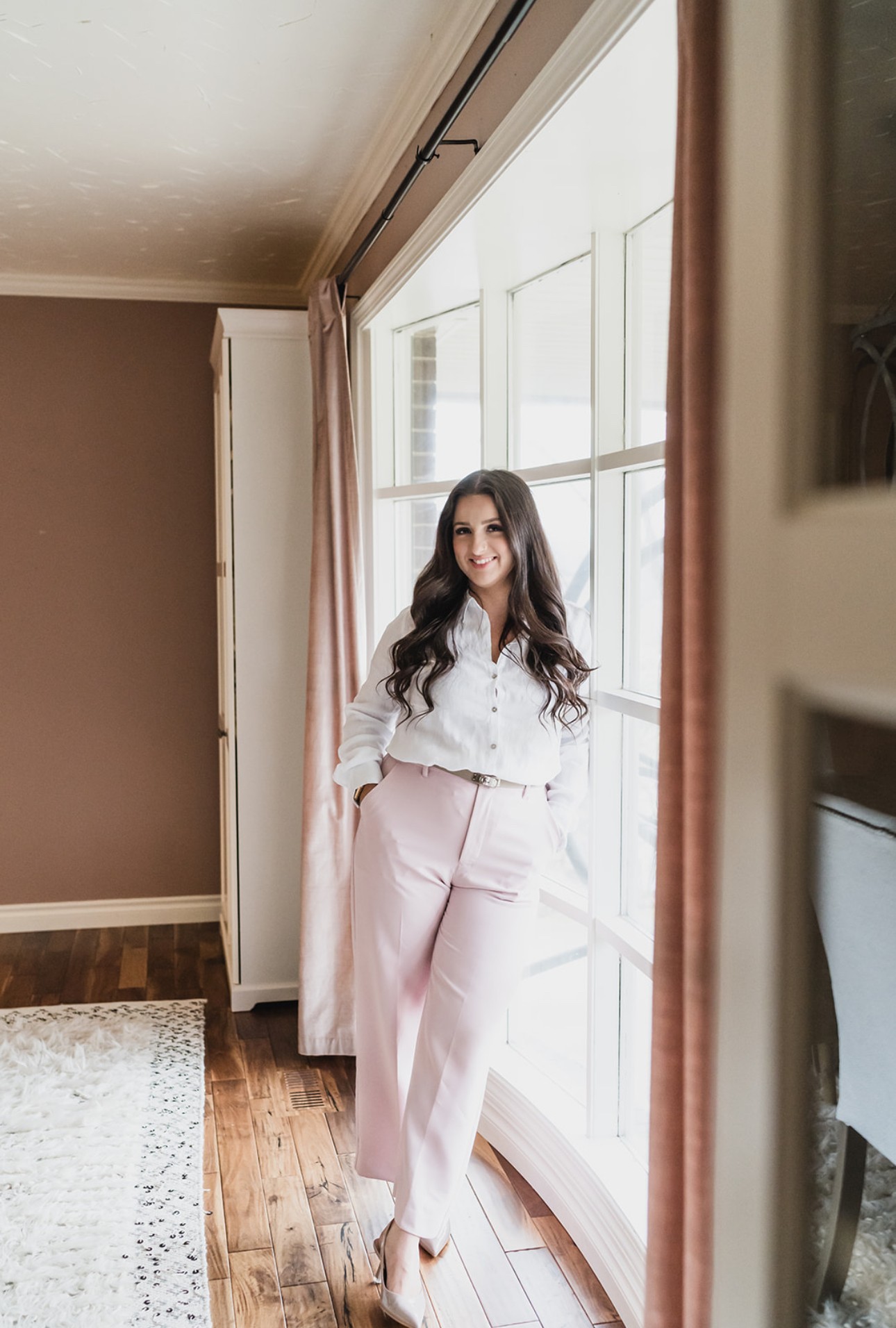 Young woman with long dark hair standing by a large window, smiling, wearing a white blouse and pink pants.
