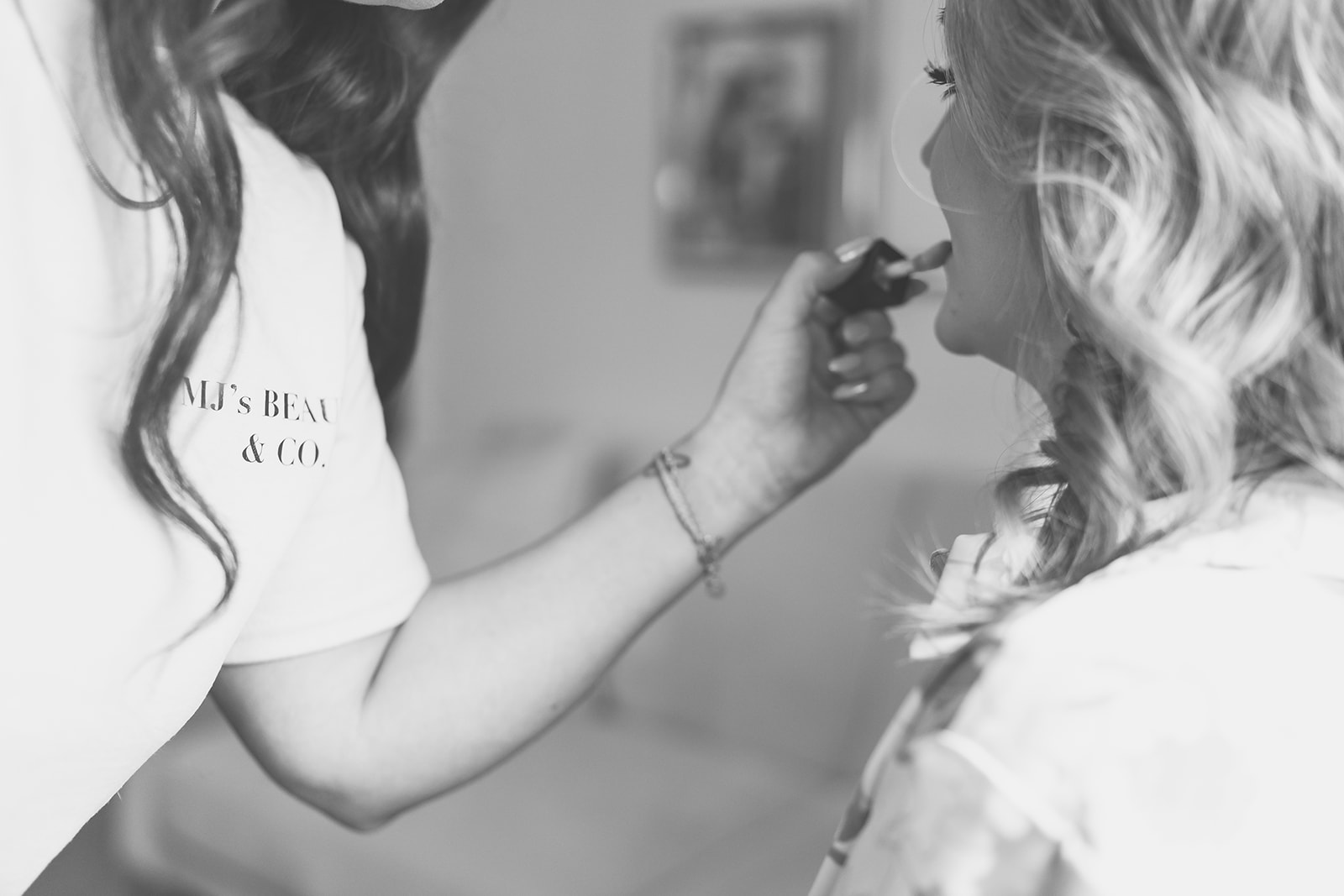 A person applies makeup to another person's lips, with a blurred background and visible hair and arm details.