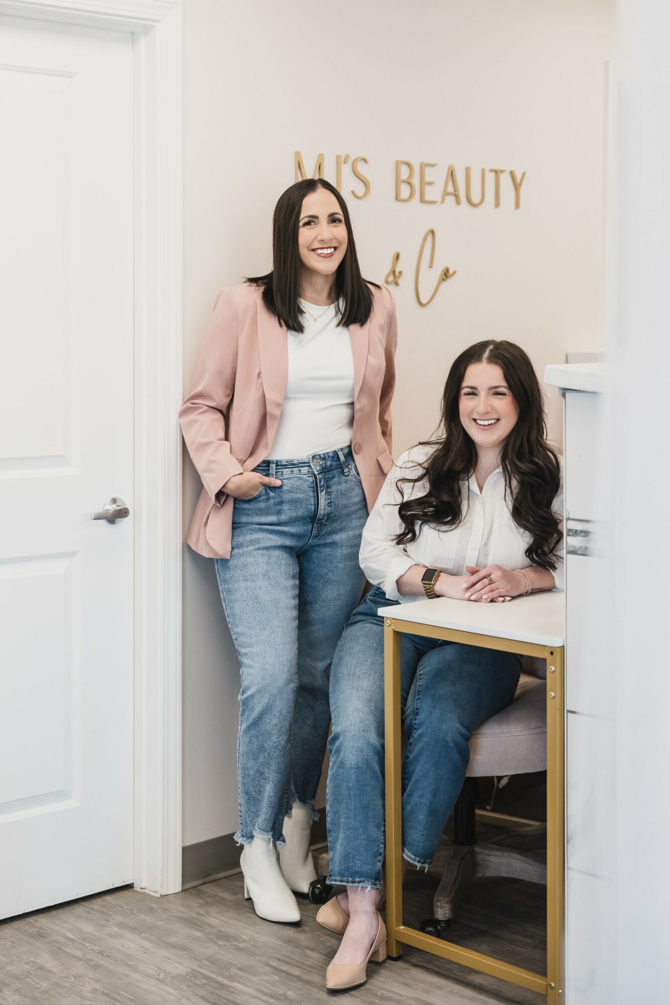 Two women in a beauty salon, one standing and one sitting at a desk, smiling, with a sign reading 'K's Beauty' behind them.