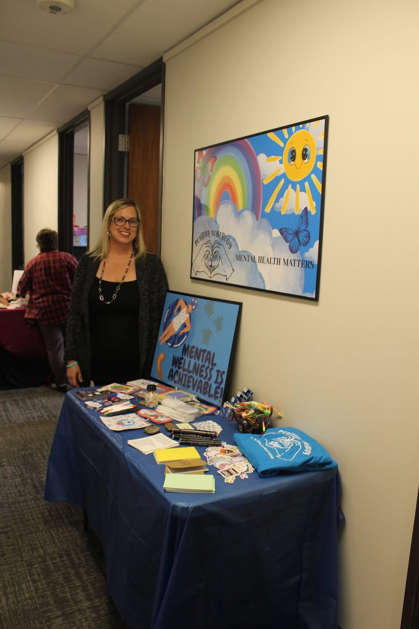 Woman standing next to a table with books and items, colorful rainbow and sun artwork on the wall behind her.