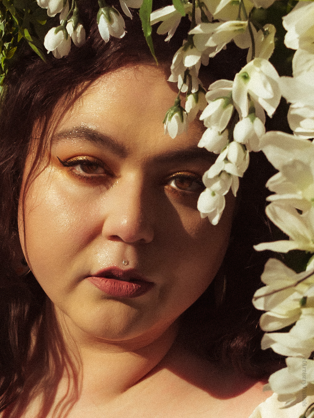 Close-up of a woman with dark hair and makeup, surrounded by white flowers, looking at the camera.
