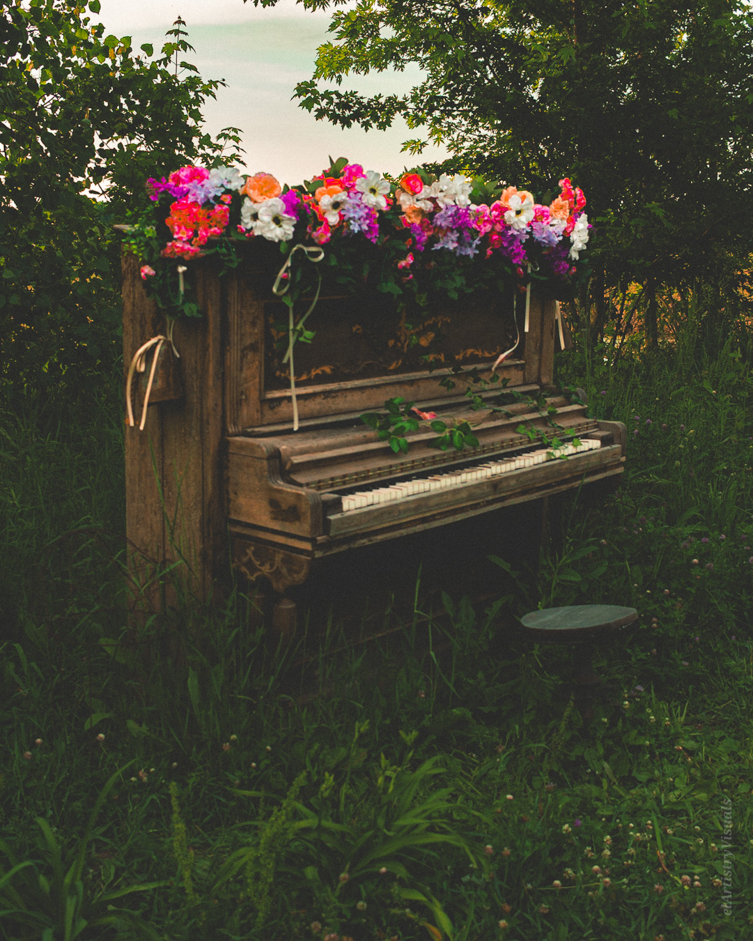 Old upright piano with flowers on top, outdoors among trees and grass, in natural light.