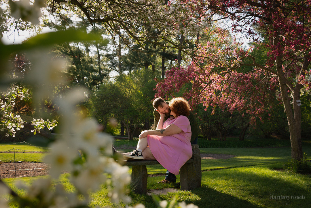 Two women sit on a bench in a park with blooming trees, one in a pink dress and the other in dark clothing, embracing.