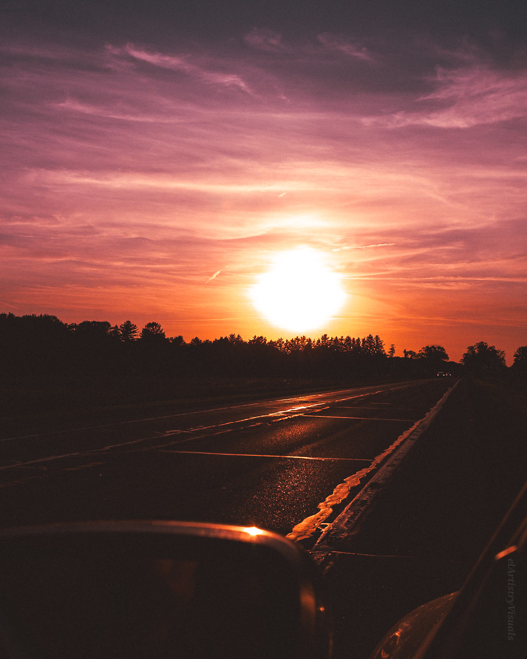 Sunset over a road with colorful sky and silhouetted trees, bright sun near horizon, reflecting on wet pavement.