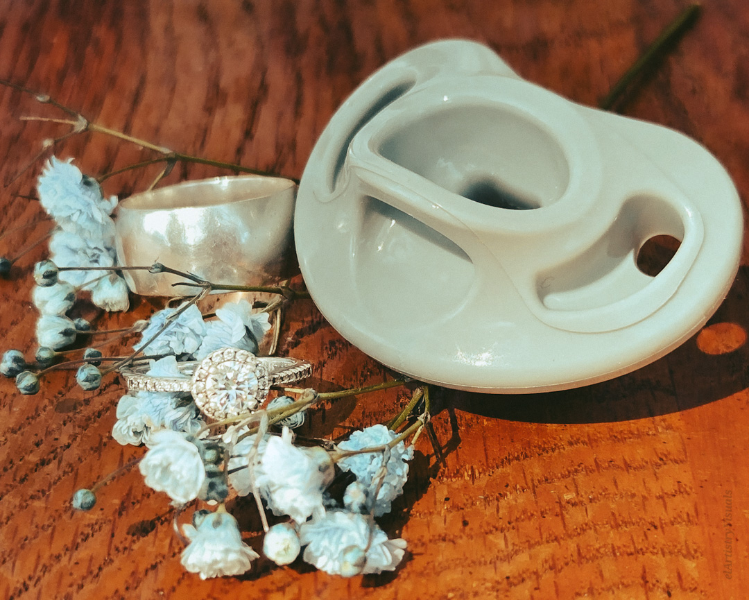 A white ceramic baby pacifier, a ring with a large gemstone, and dried blue flowers on a wooden surface.