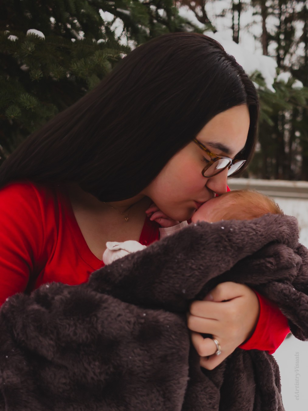 Woman with glasses and long dark hair holding a newborn wrapped in a dark blanket outdoors.