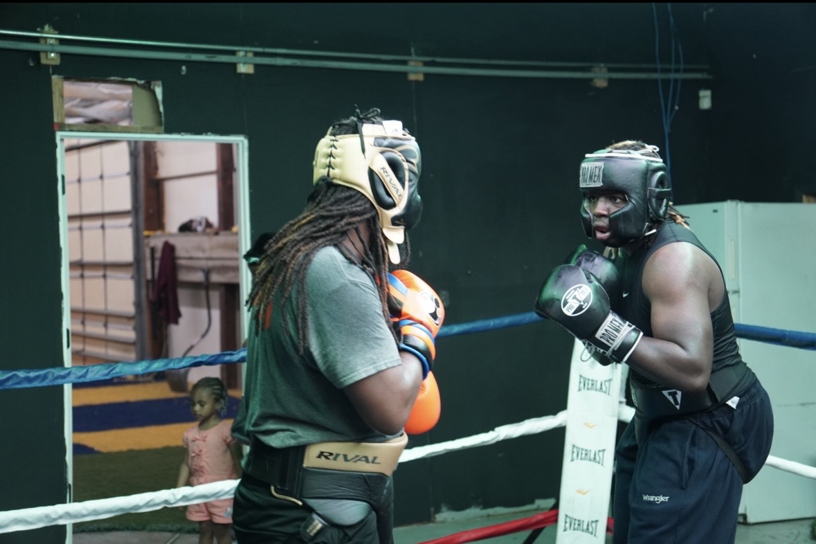 Two women in boxing gear face each other in a boxing ring, one with dreadlocks and the other with a helmet, gloves, and sleeveless shirt.