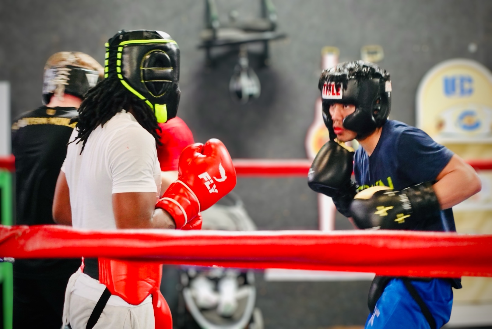 Two young people in boxing gear face each other in a boxing ring, wearing helmets and gloves, with a red rope barrier.