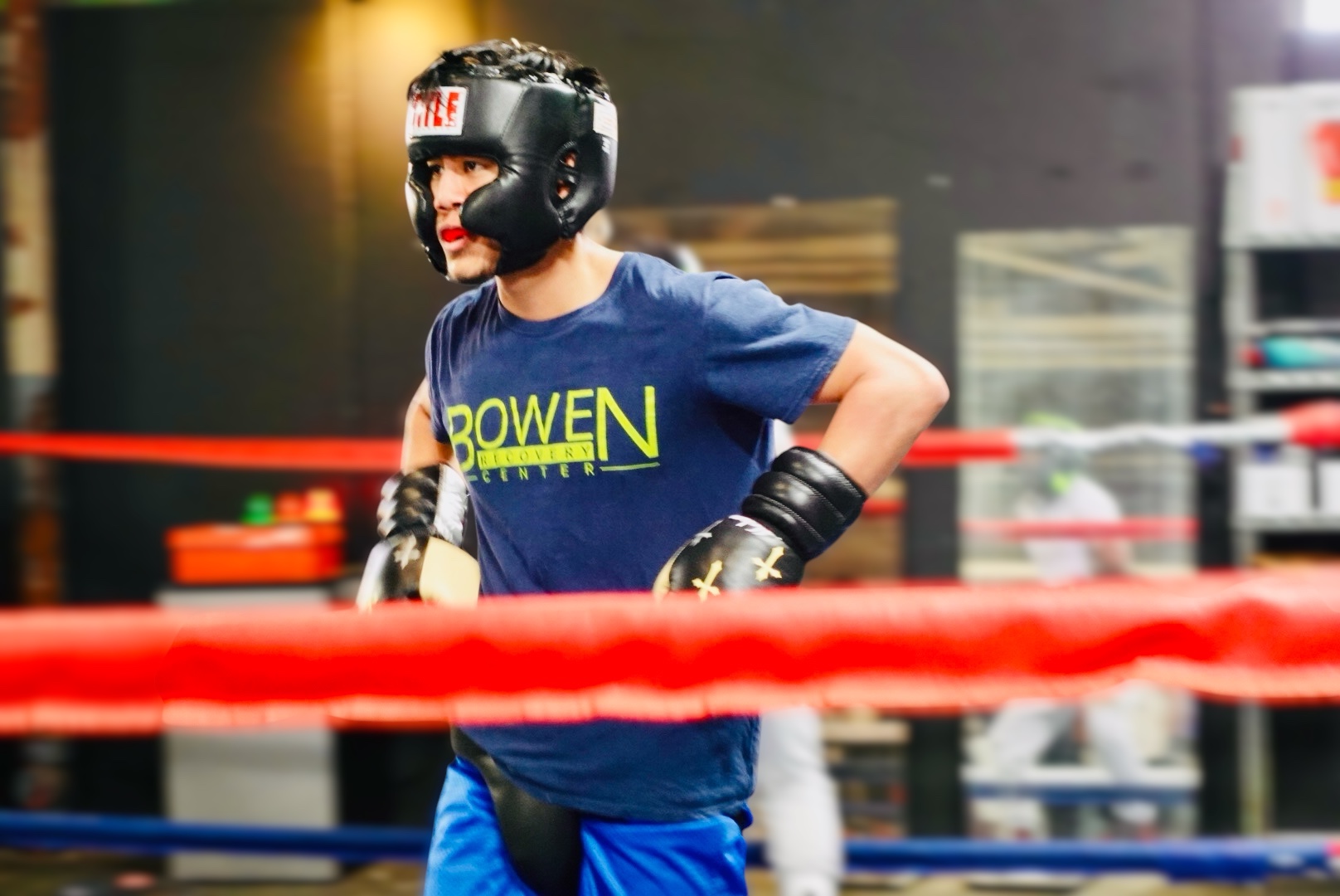 Person wearing a helmet and gloves, standing in a boxing ring with red and blue ropes, in an indoor gym.
