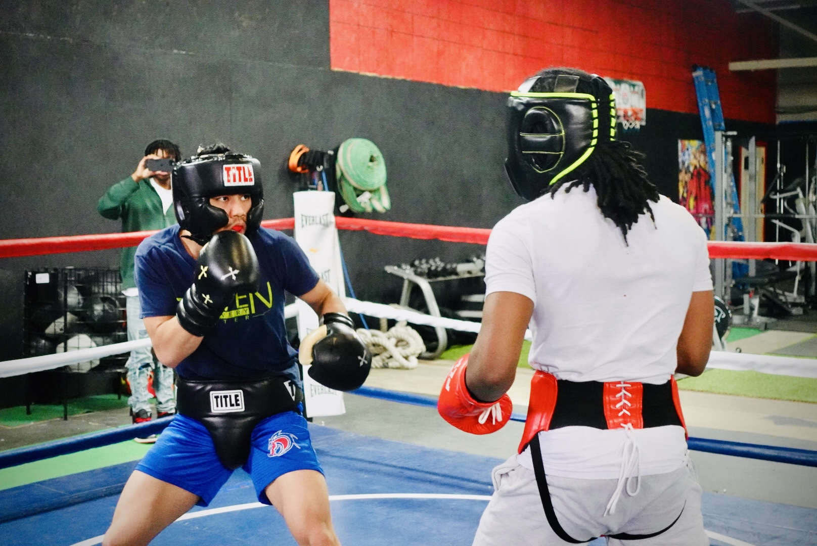 Two people in boxing gear face each other in a boxing ring, one in blue and the other in white, with protective headgear.