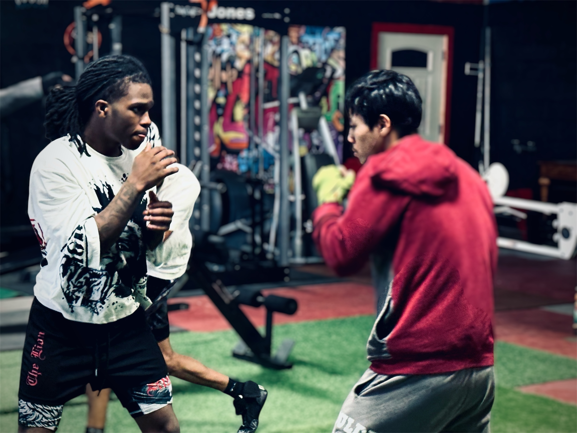 Two people in a gym, one with dreadlocks and a white shirt, the other in a red hoodie, facing each other.