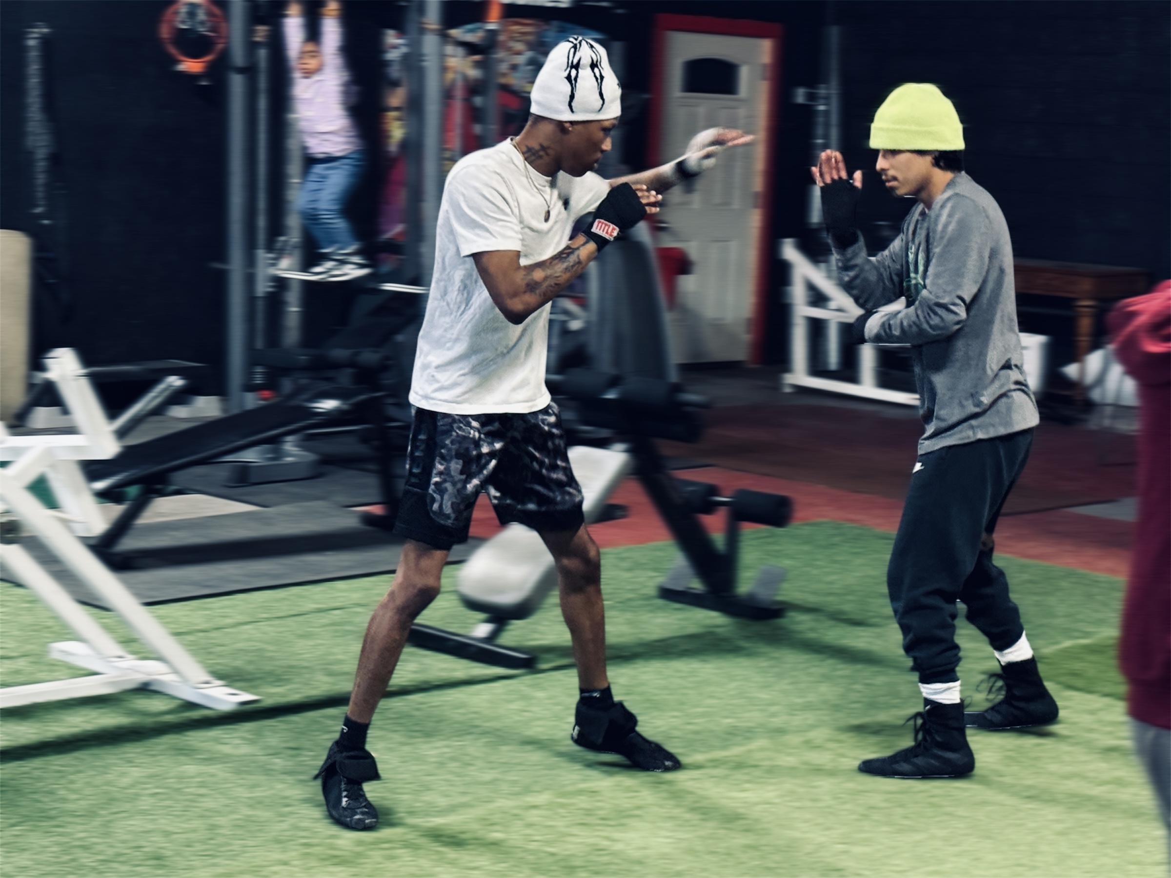 Two people training with a punching bag in a gym, surrounded by workout equipment, on a green floor.