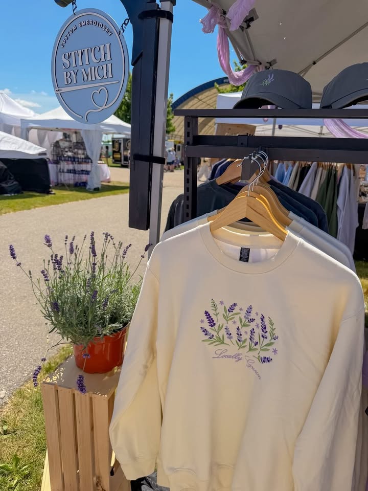 Cream-colored sweatshirt with purple floral design hanging on a rack outdoors, with lavender plant in foreground and market tents in background.