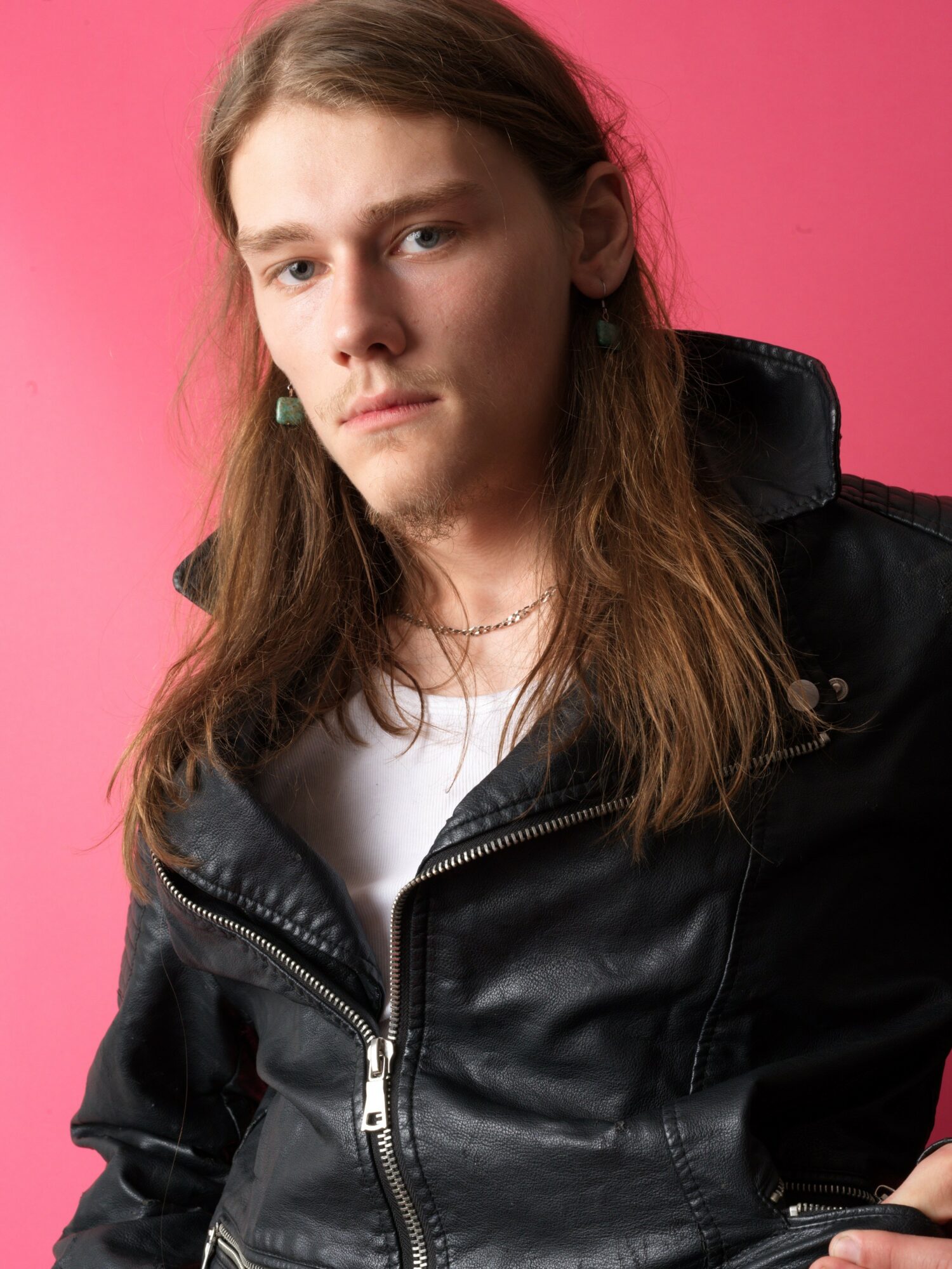 Young person with long hair wearing a black leather jacket and white shirt, against a pink background.