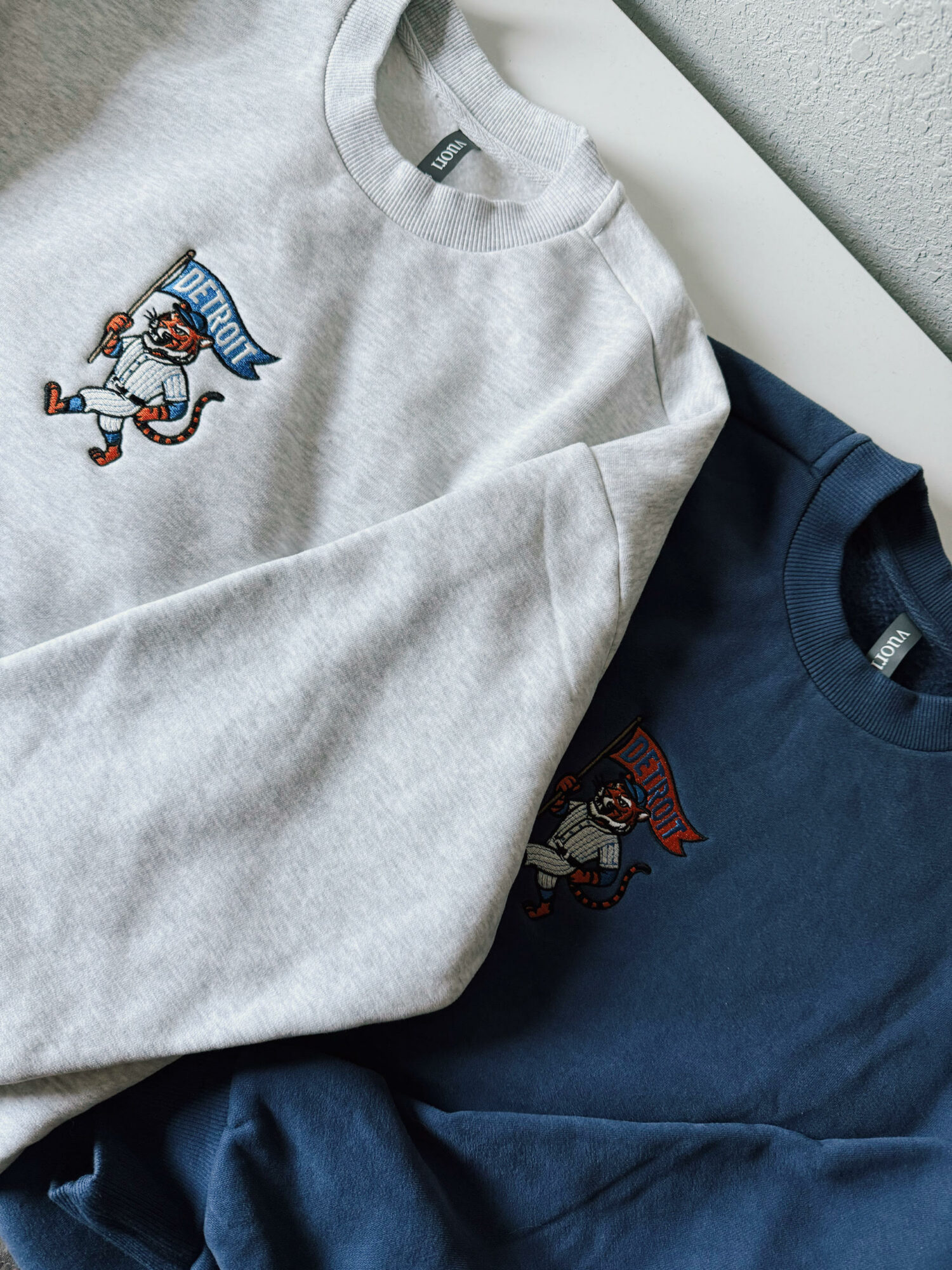 Two folded sweatshirts, one light gray with a snowman embroidery and one navy blue with a similar snowman embroidery, on a white surface.