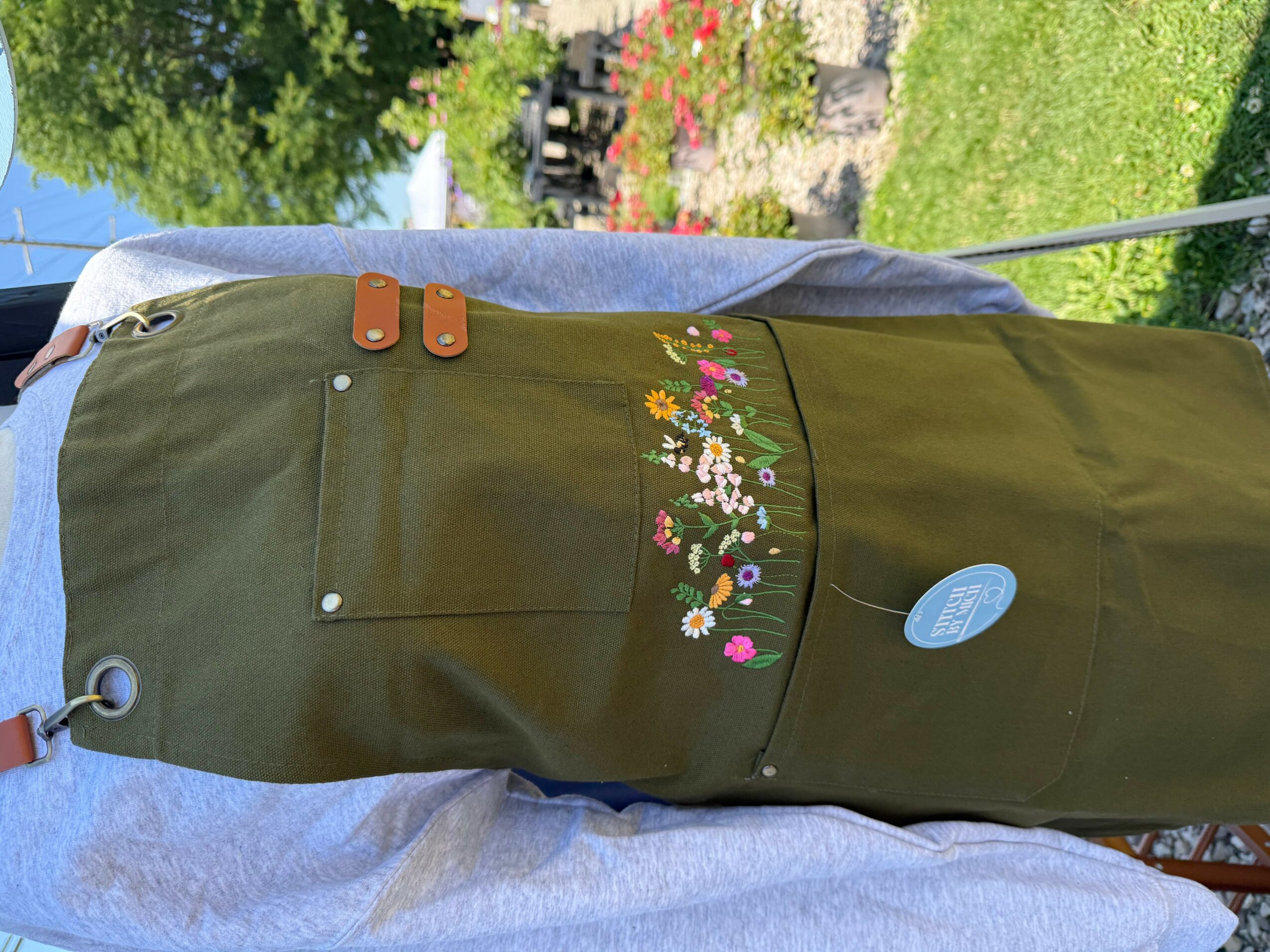 Person wearing a green apron with floral embroidery, standing outdoors with grass and flowers in the background.