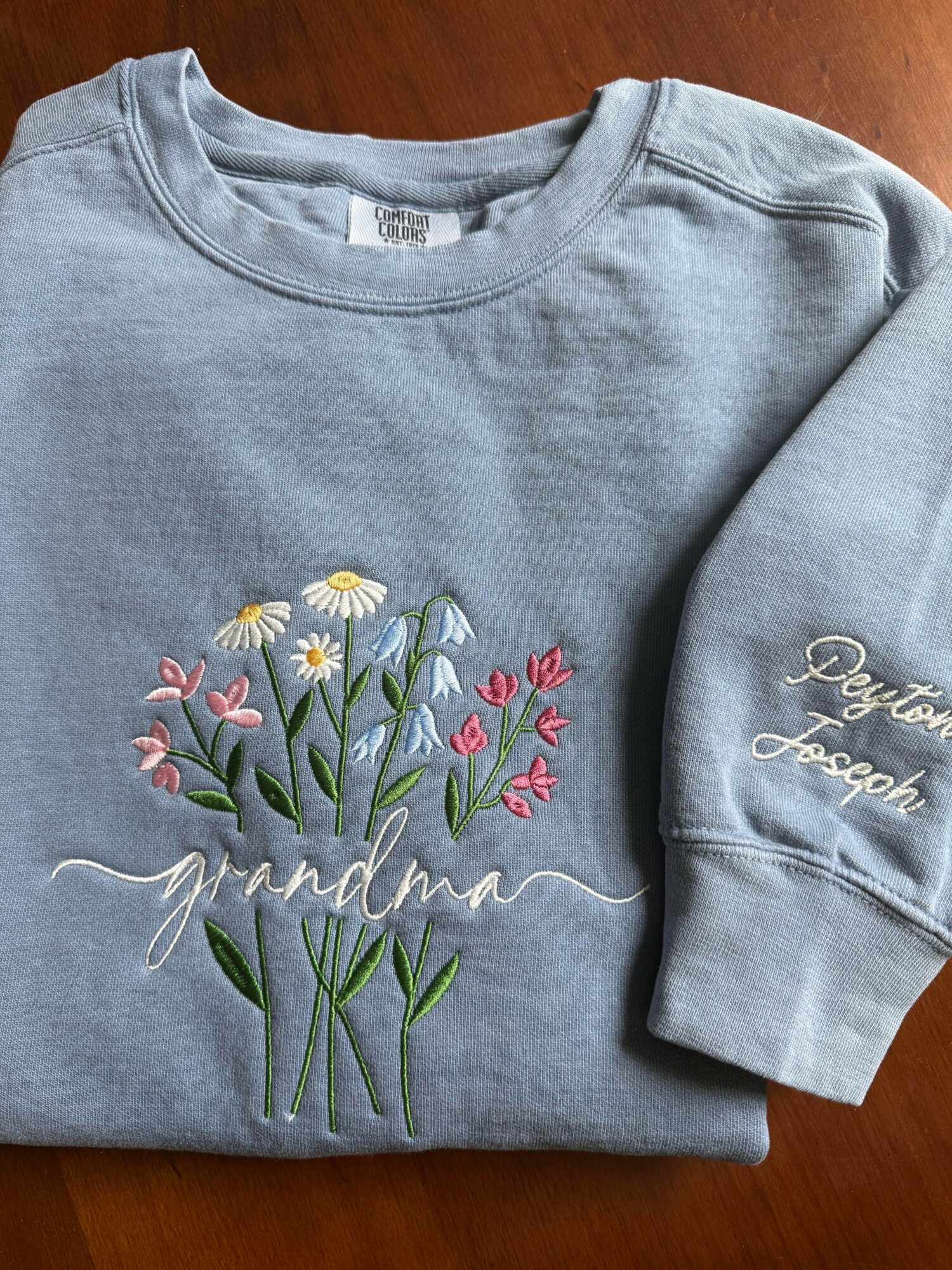 Light blue sweatshirt with embroidered daisies and tulips, and the word 'grandma' in cursive, on a wooden surface.