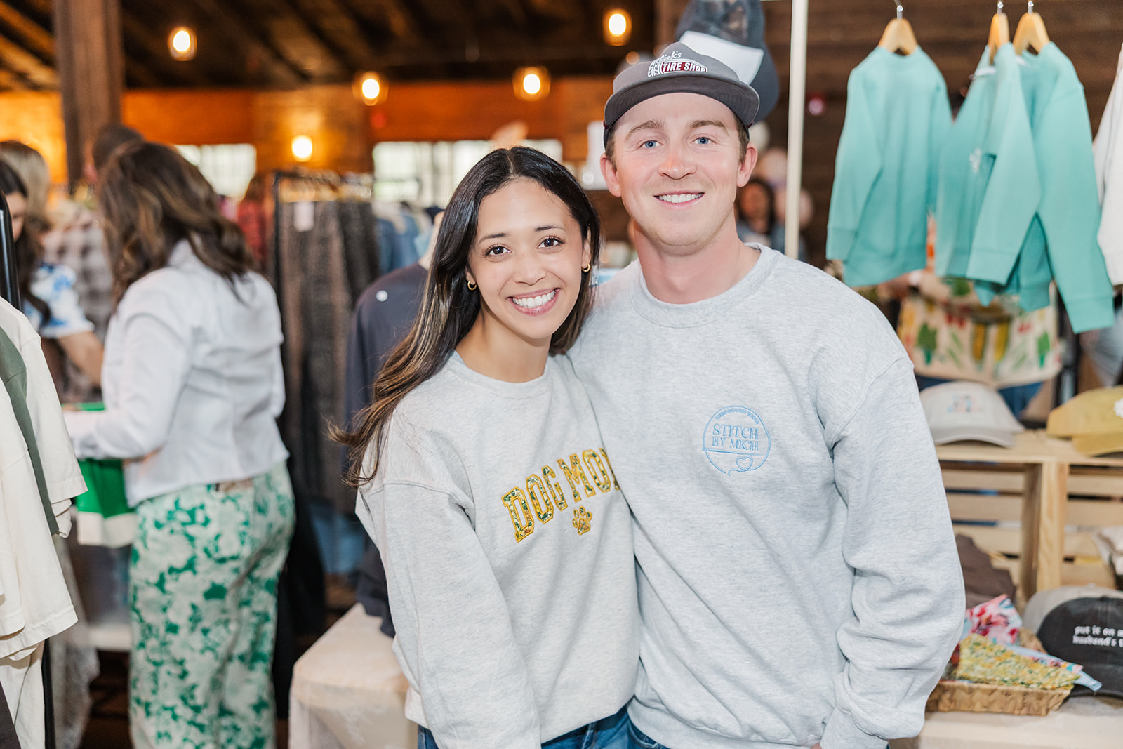 Smiling young woman and man standing together at an outdoor market, surrounded by clothing and people, smiling at the camera.