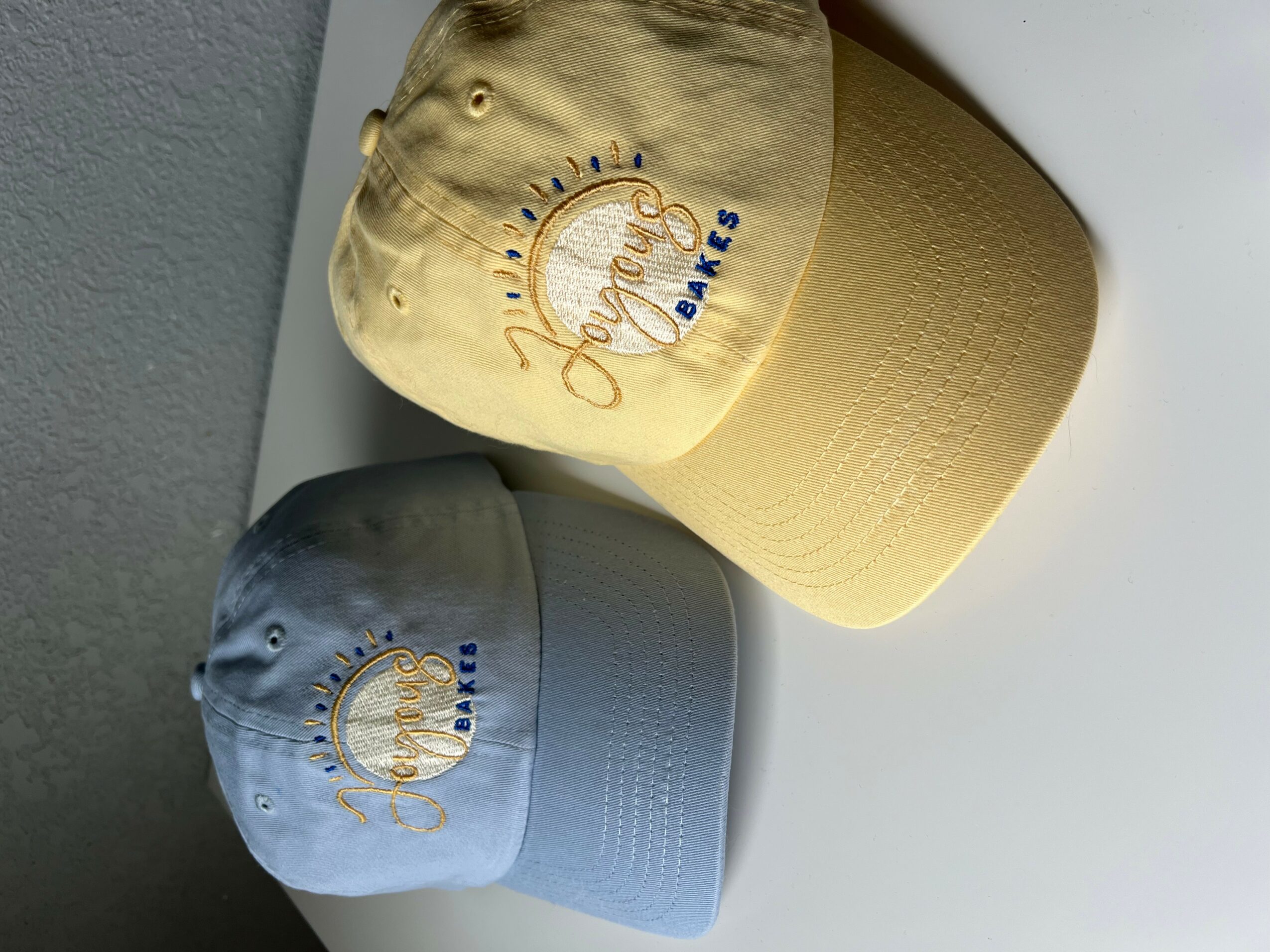Two baseball caps, one yellow and one gray, with embroidered logos, placed on a surface.