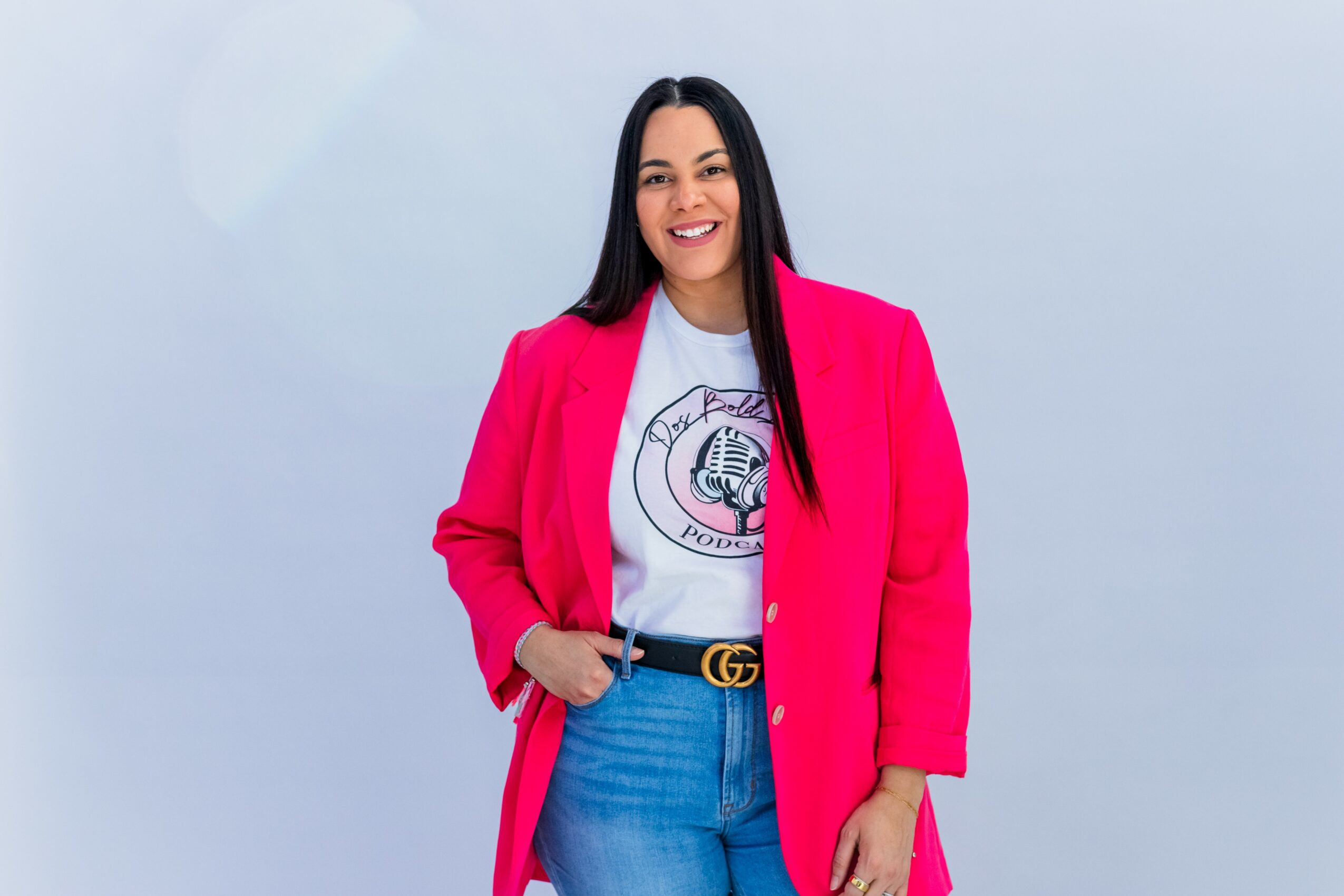 Woman with long dark hair wearing a bright pink blazer, white graphic t-shirt, and blue jeans, smiling against a light background.