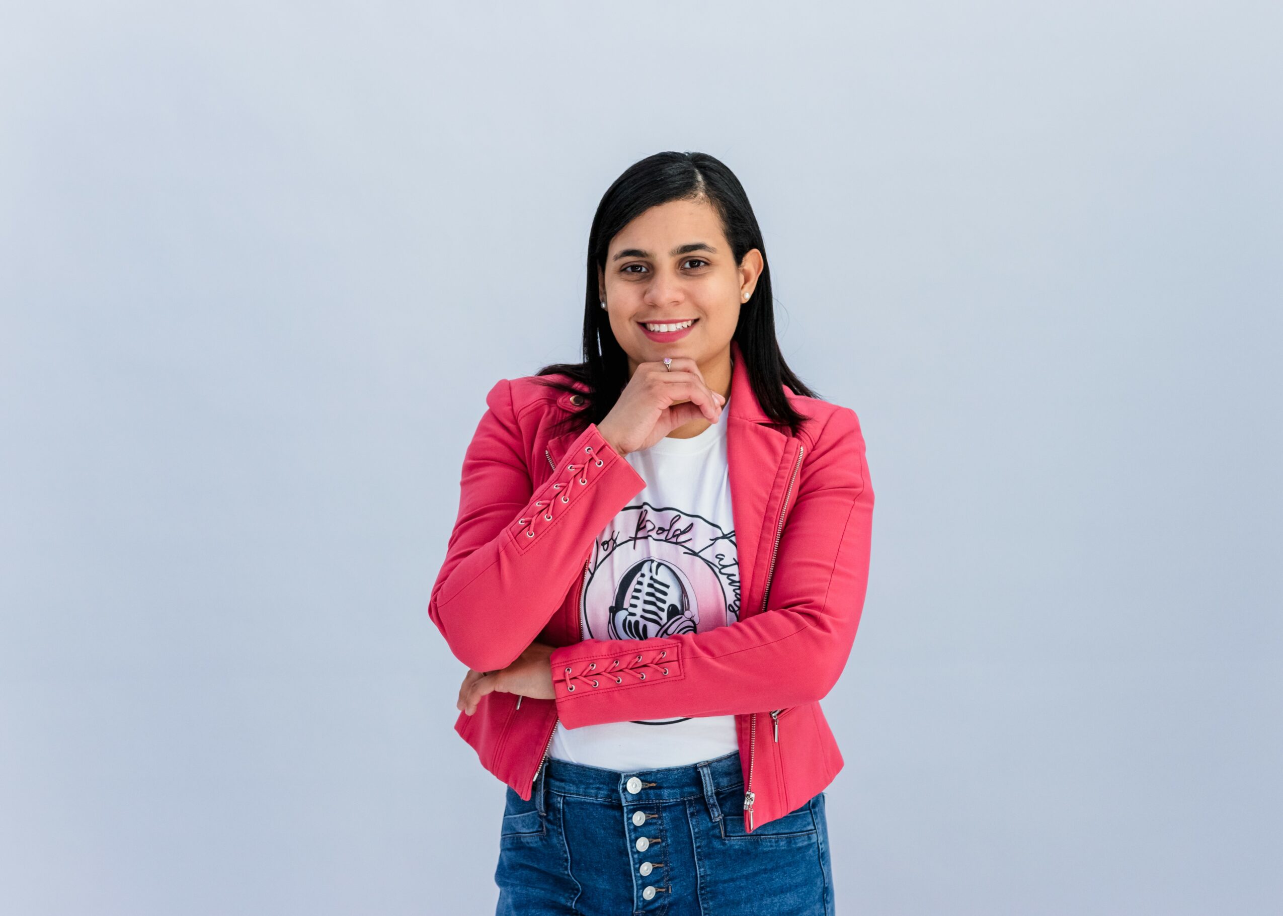 Young woman with dark hair smiling, wearing a pink jacket and white t-shirt, standing against a light blue background.