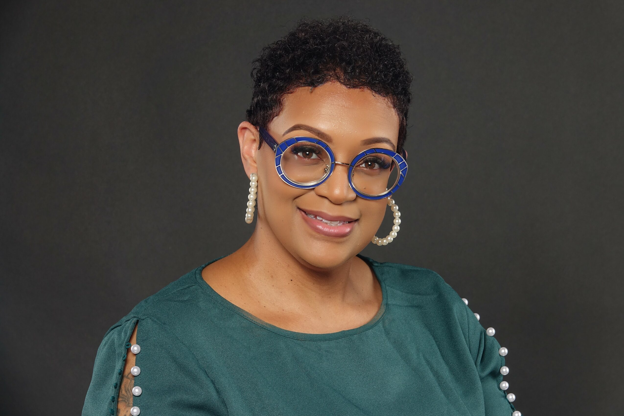 Portrait of a woman with short curly hair, wearing large round glasses, pearl earrings, and a green top with pearl embellishments on the shoulder.