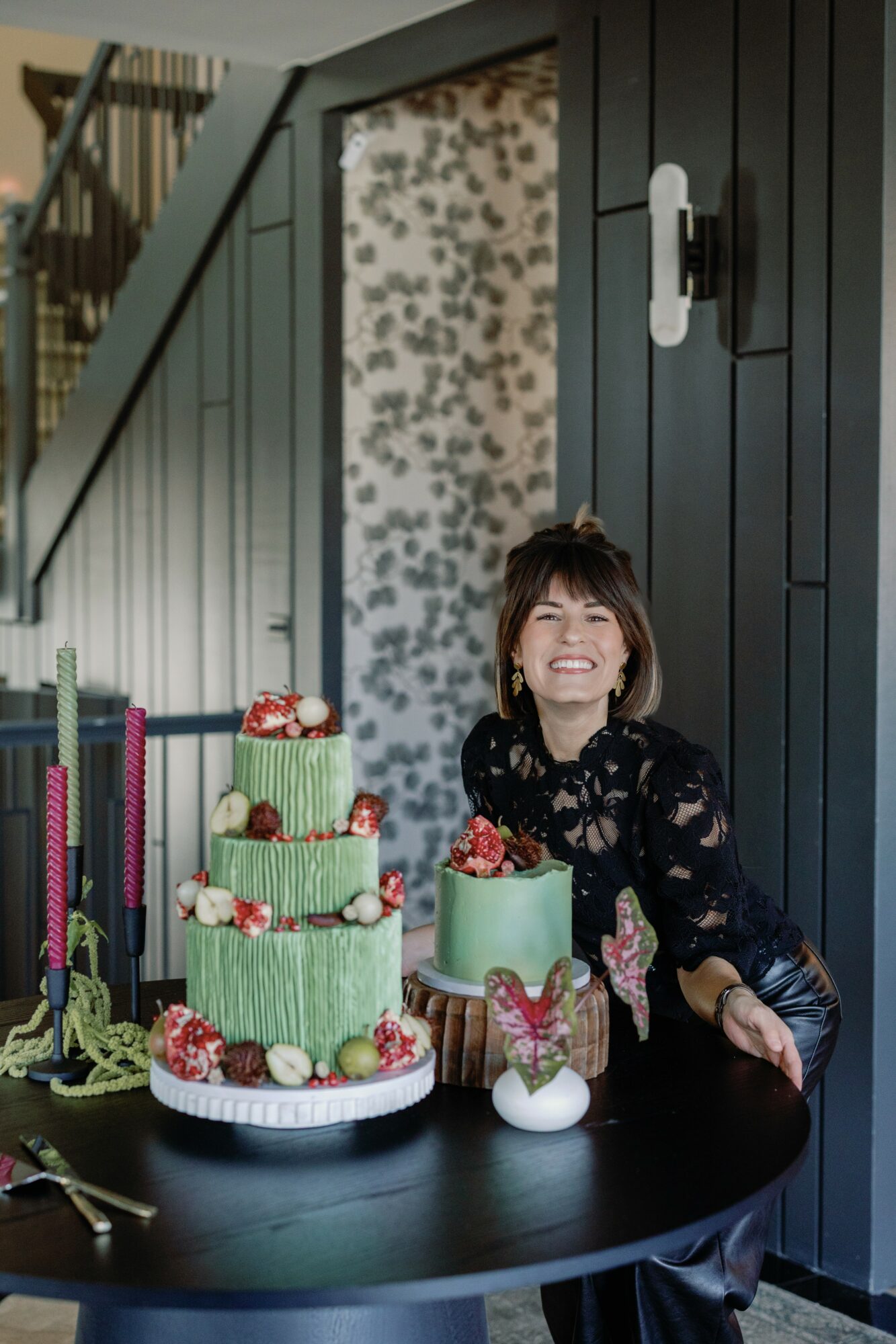 Woman smiling at a table with three decorated cakes, in a room with dark paneling and patterned wallpaper.