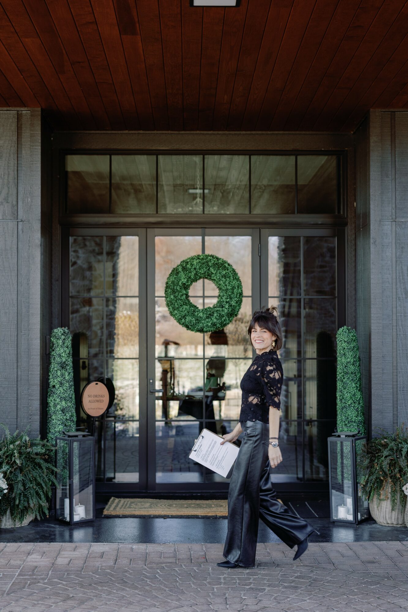 Woman standing outside glass door with a wreath, holding a paper, surrounded by potted plants, in front of a building.