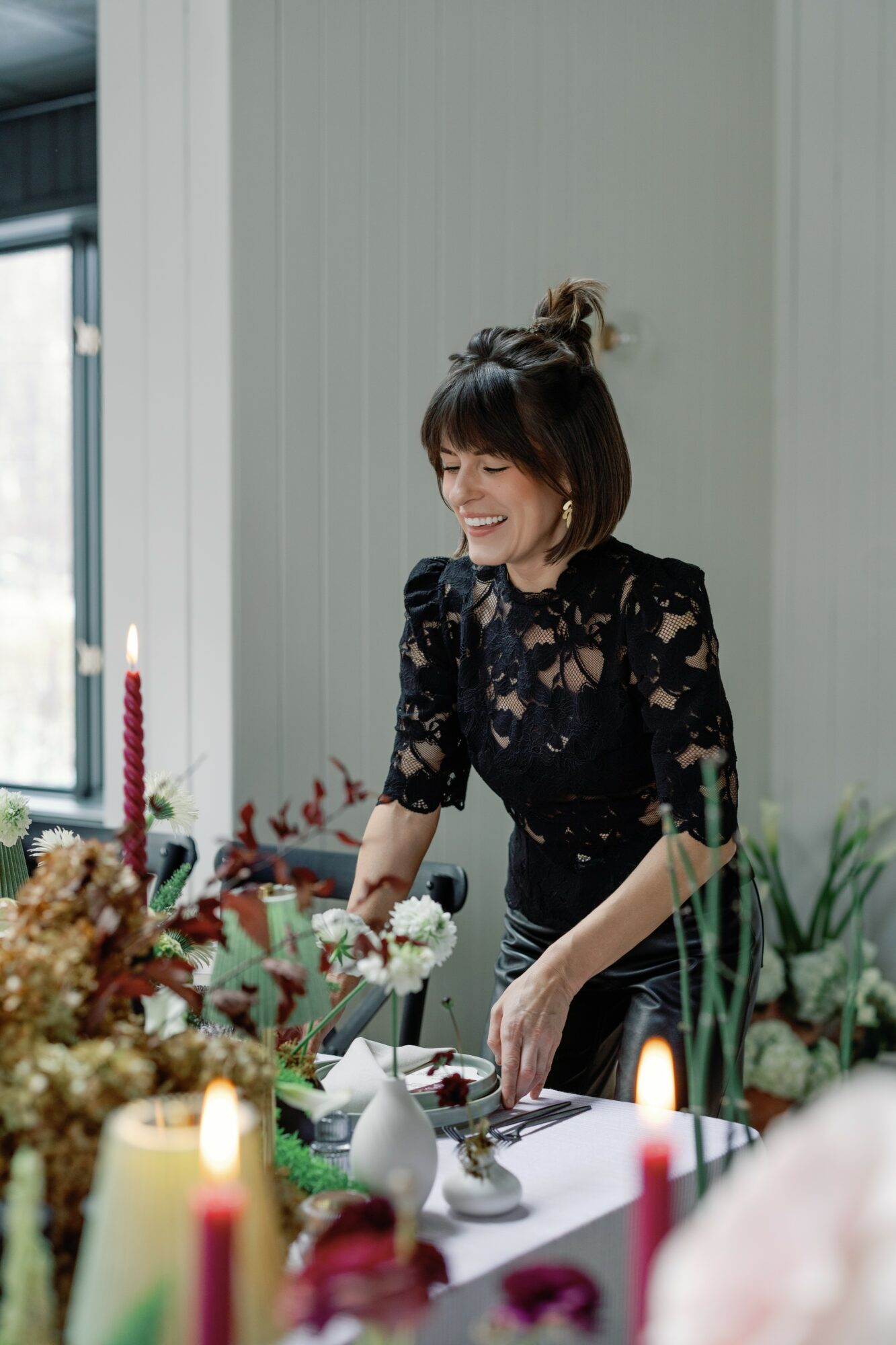 Woman with short dark hair and a black lace dress arranging flowers at a table, smiling, with candles and plants nearby.