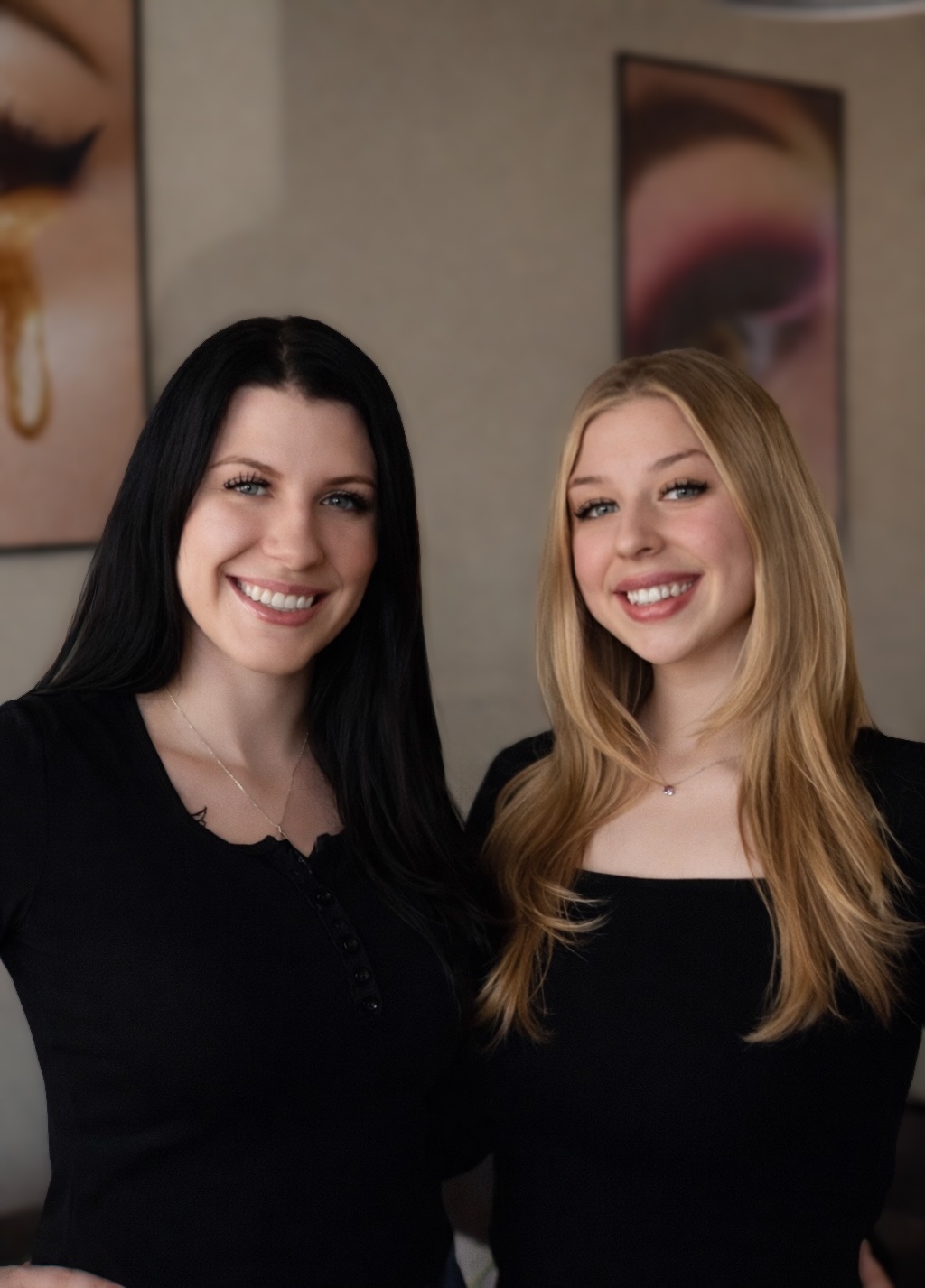 Two women smiling, standing close together indoors, with artwork on the wall behind them.