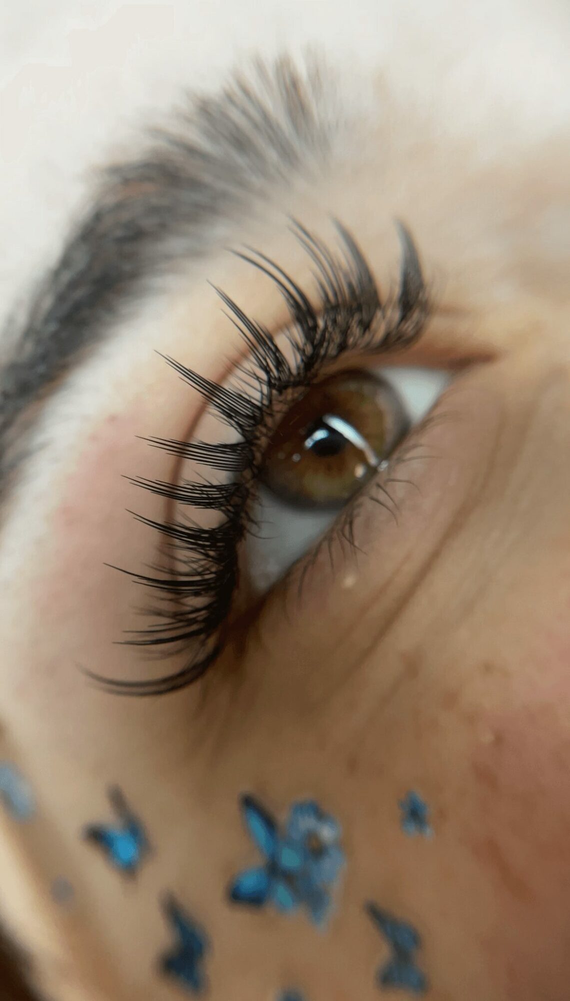 Close-up of a person's eye with long, curled eyelashes and brown iris, with blue decorative elements below eye.