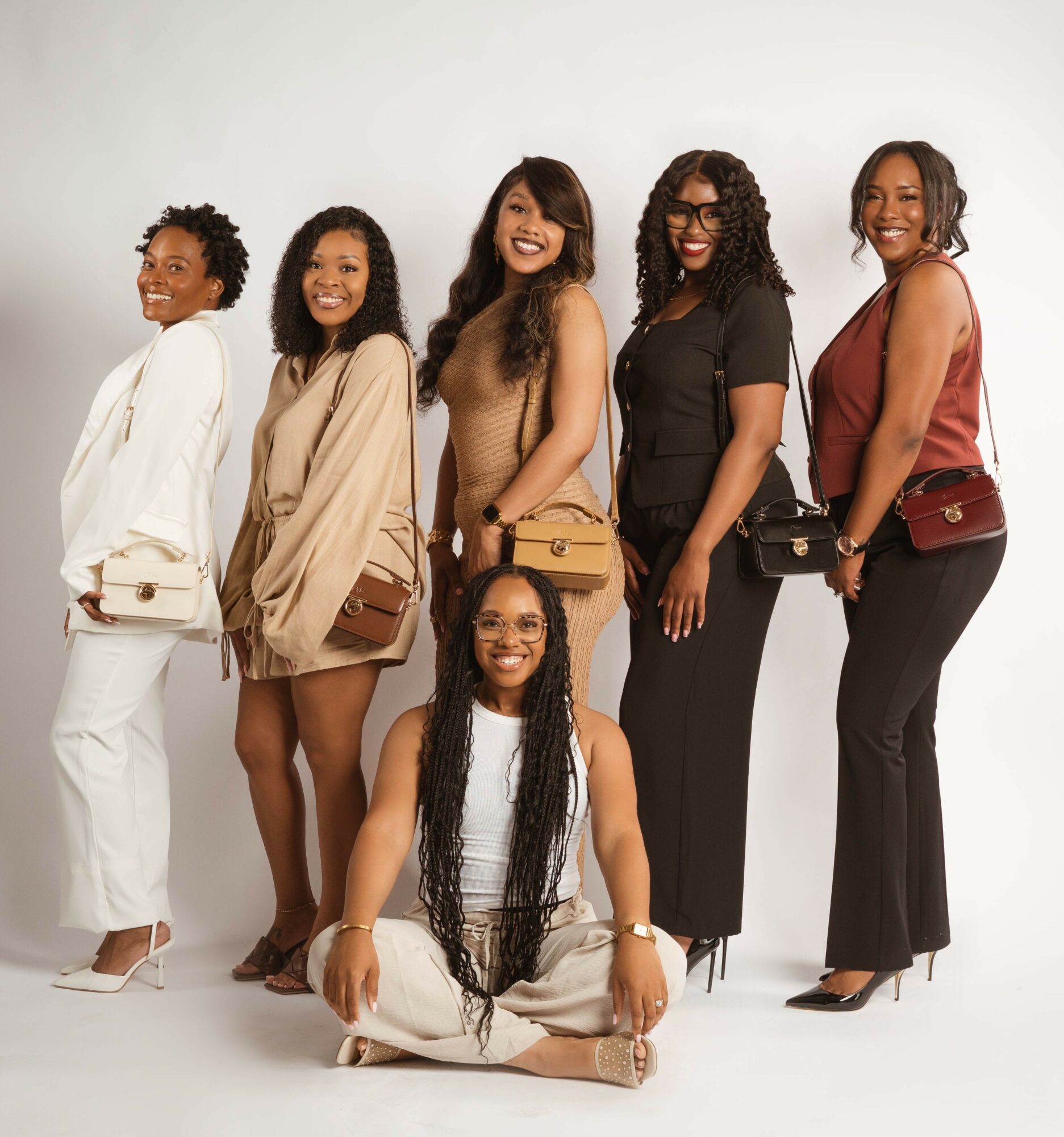 Group of six women smiling, standing and sitting against a plain background, dressed in stylish casual and formal clothing.