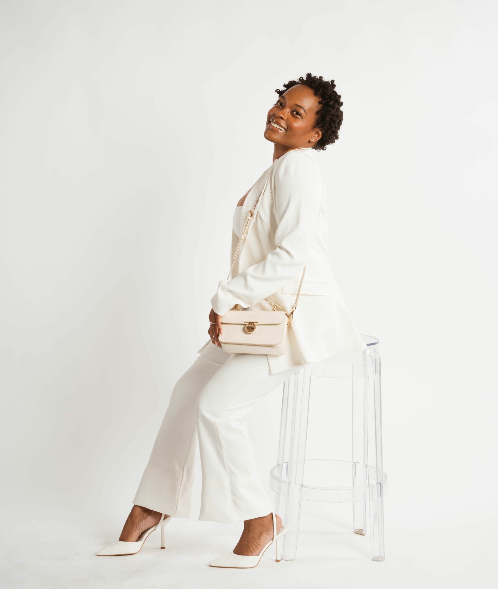 Woman sitting on a transparent chair, smiling, wearing a white outfit, holding a beige purse, against a plain white background.