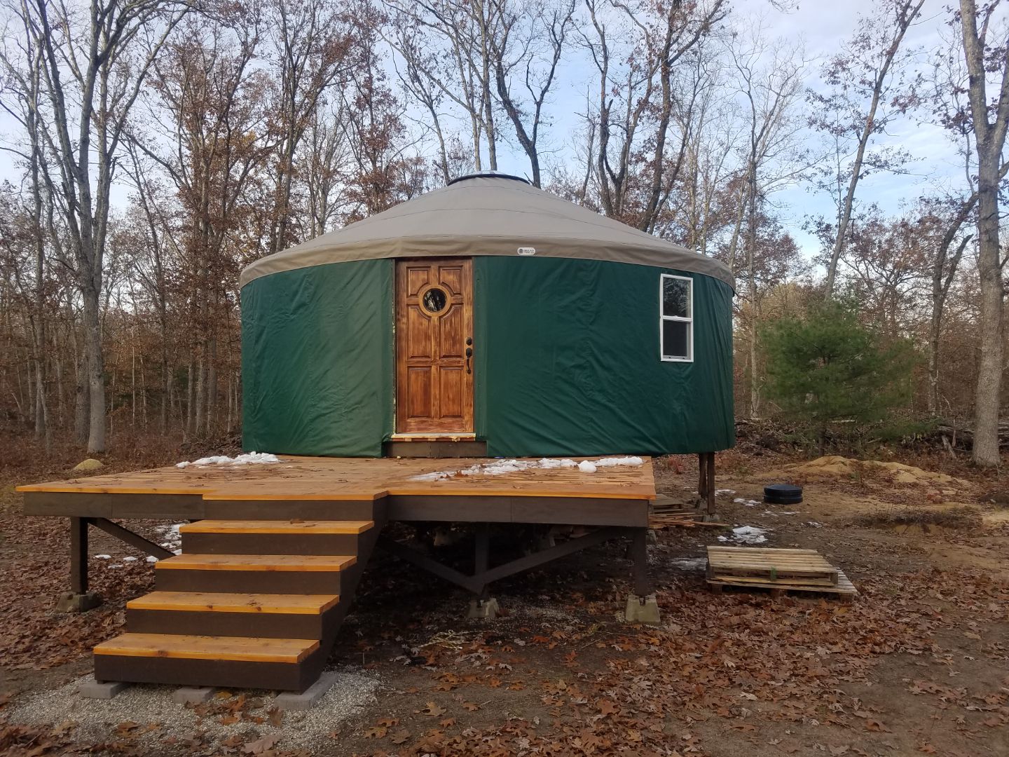 A round, green yurt with a wooden door, elevated on a wooden platform with stairs, outdoors with trees in background.