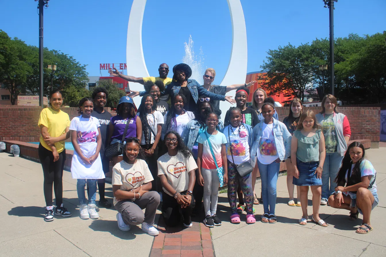 Group of people posing outdoors in front of a large white sculpture, with trees and a blue sky in the background.