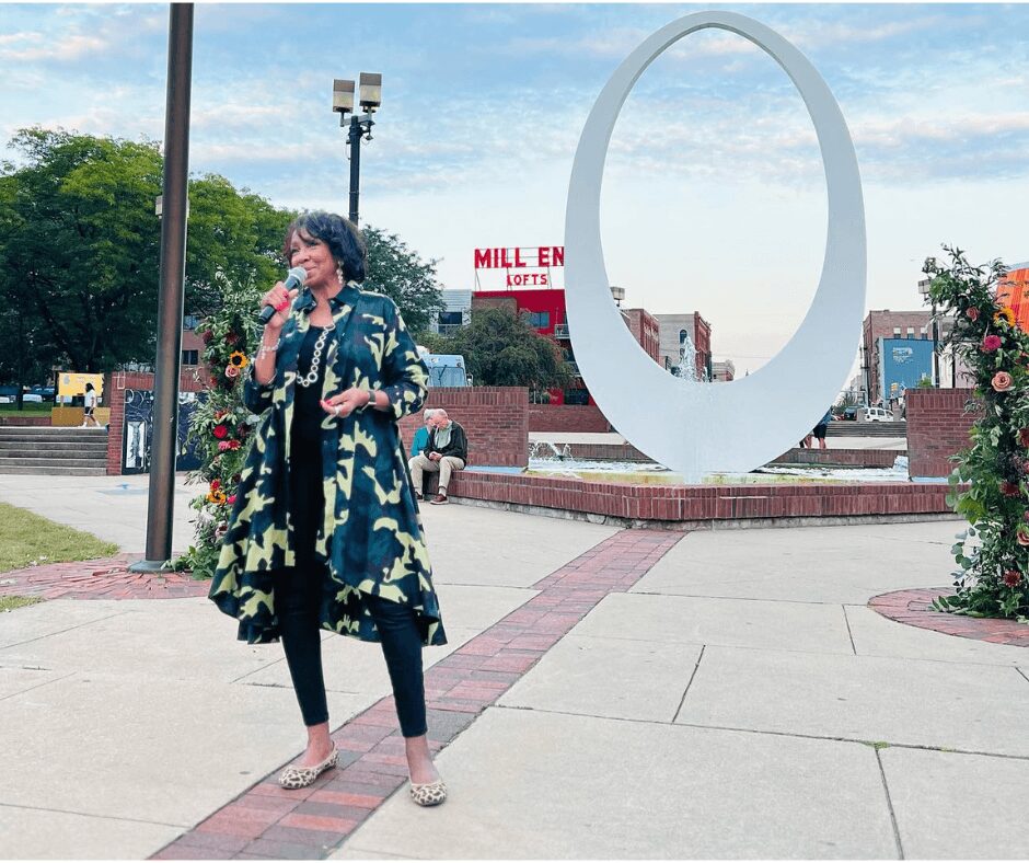 Woman speaking into microphone outdoors near large circular sculpture, with trees and buildings in background.