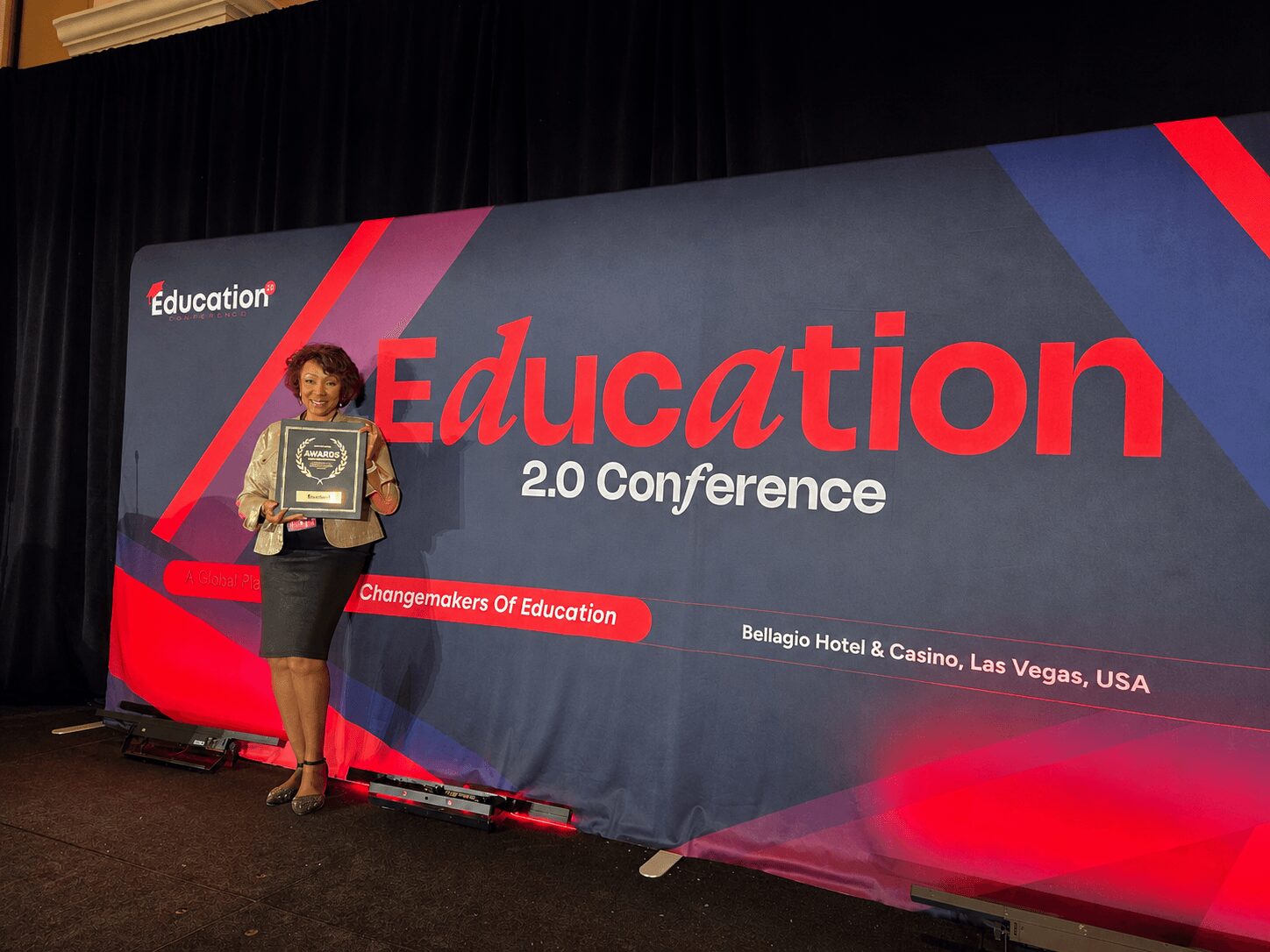 Woman holding an award standing on stage in front of a large conference banner that reads 'Education 2.0 Conference' in red and white text.