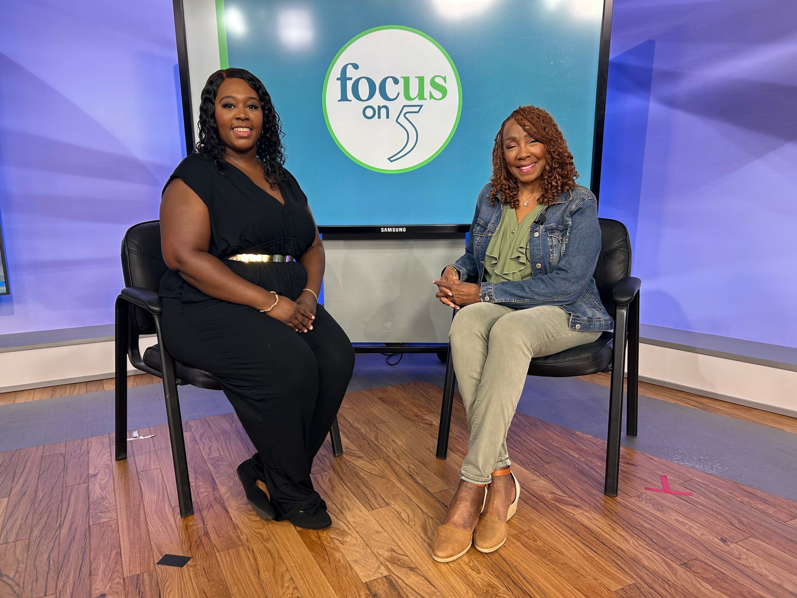 Two women sit on chairs in a TV studio with a screen behind them displaying 'focus on 5'.