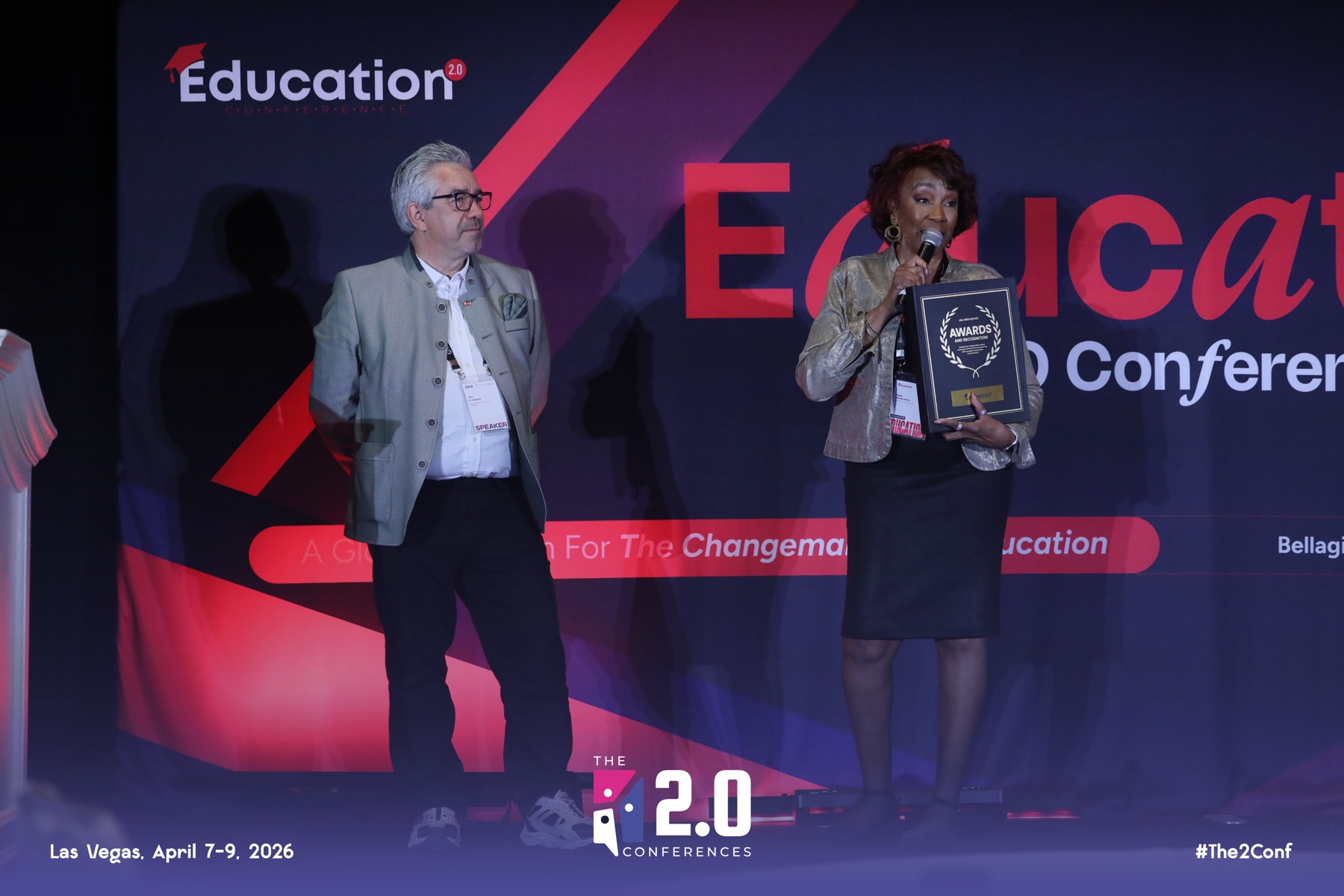Two people on stage at a conference, one woman holding an award, a man standing nearby, dark background with conference branding.