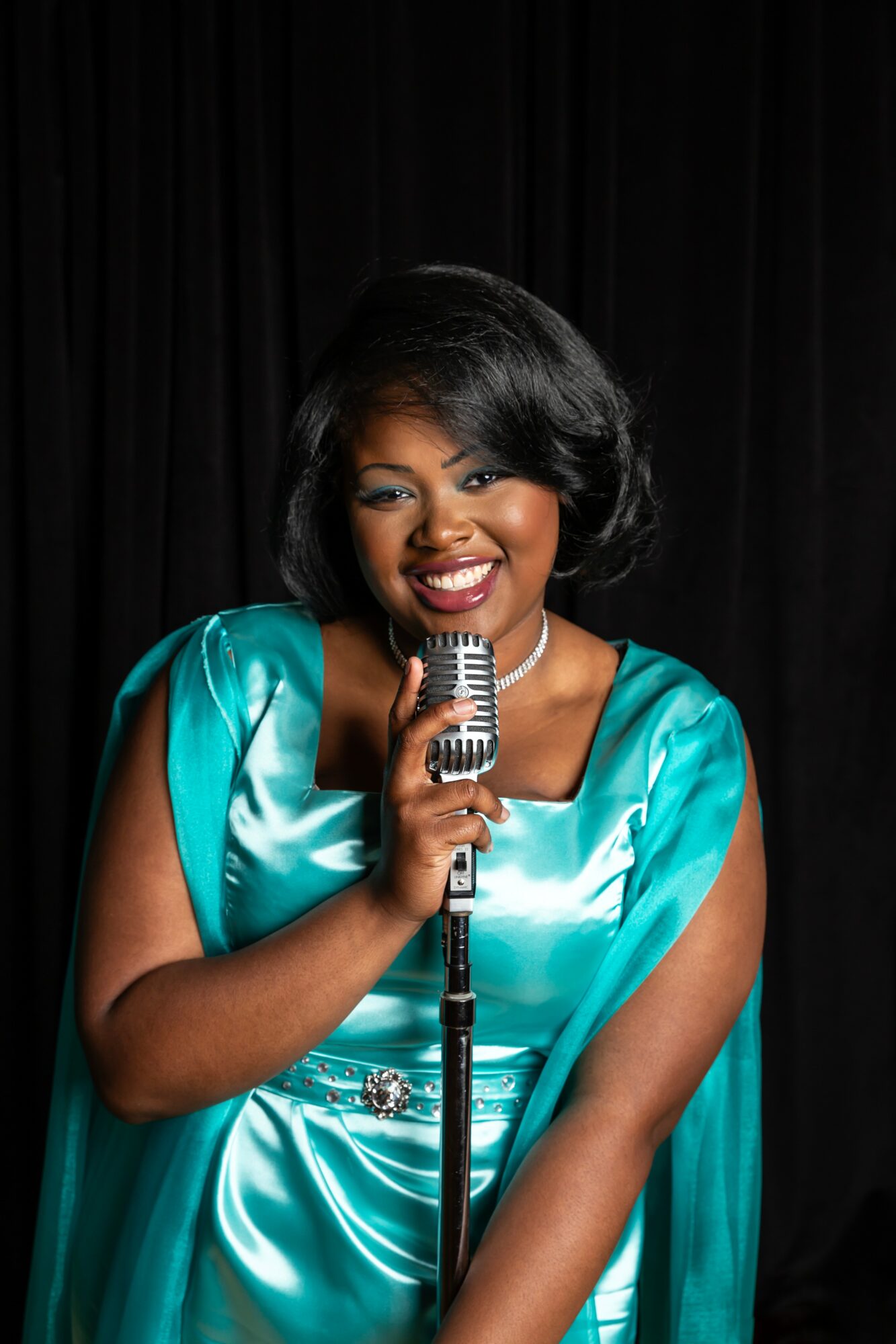 Woman in teal dress holding a vintage microphone, smiling, black background, studio setting.