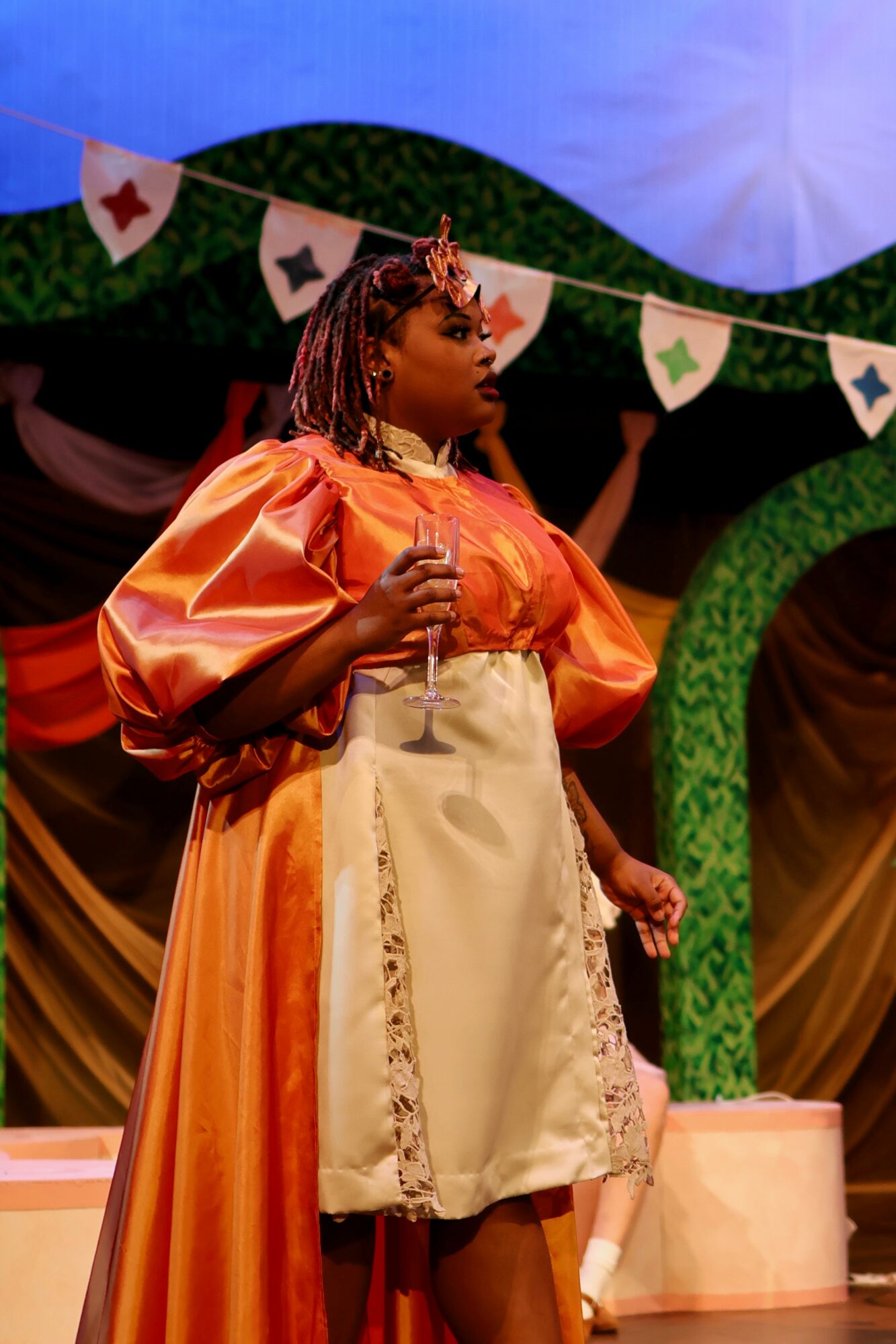 Young woman in orange and cream dress holding a microphone on stage with colorful decorations.