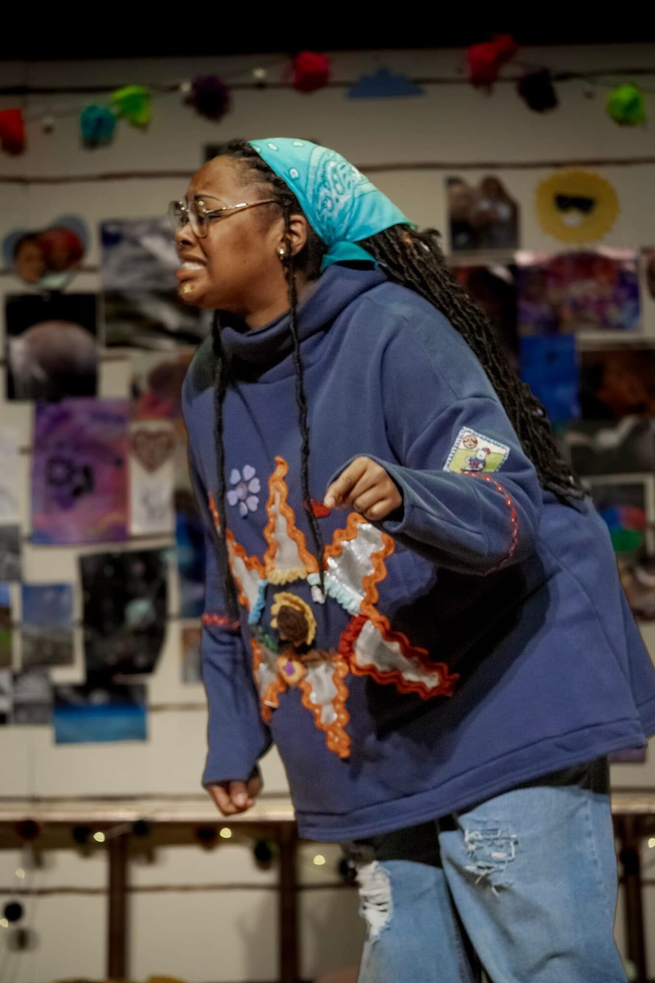 Woman with glasses, blue headscarf, and hoodie, smiling, holding a colorful star-shaped craft, in a room with photos on the wall.