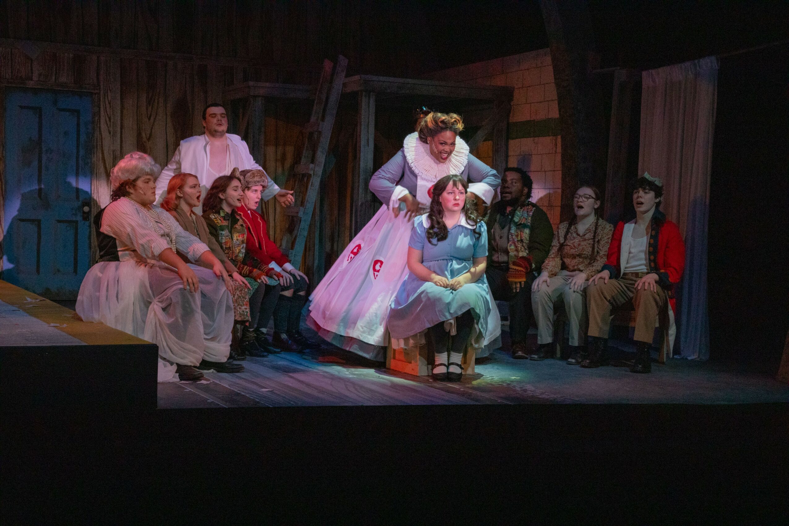 People on stage in a theatrical scene, some sitting and some standing, with a woman in a pink dress interacting with a girl.