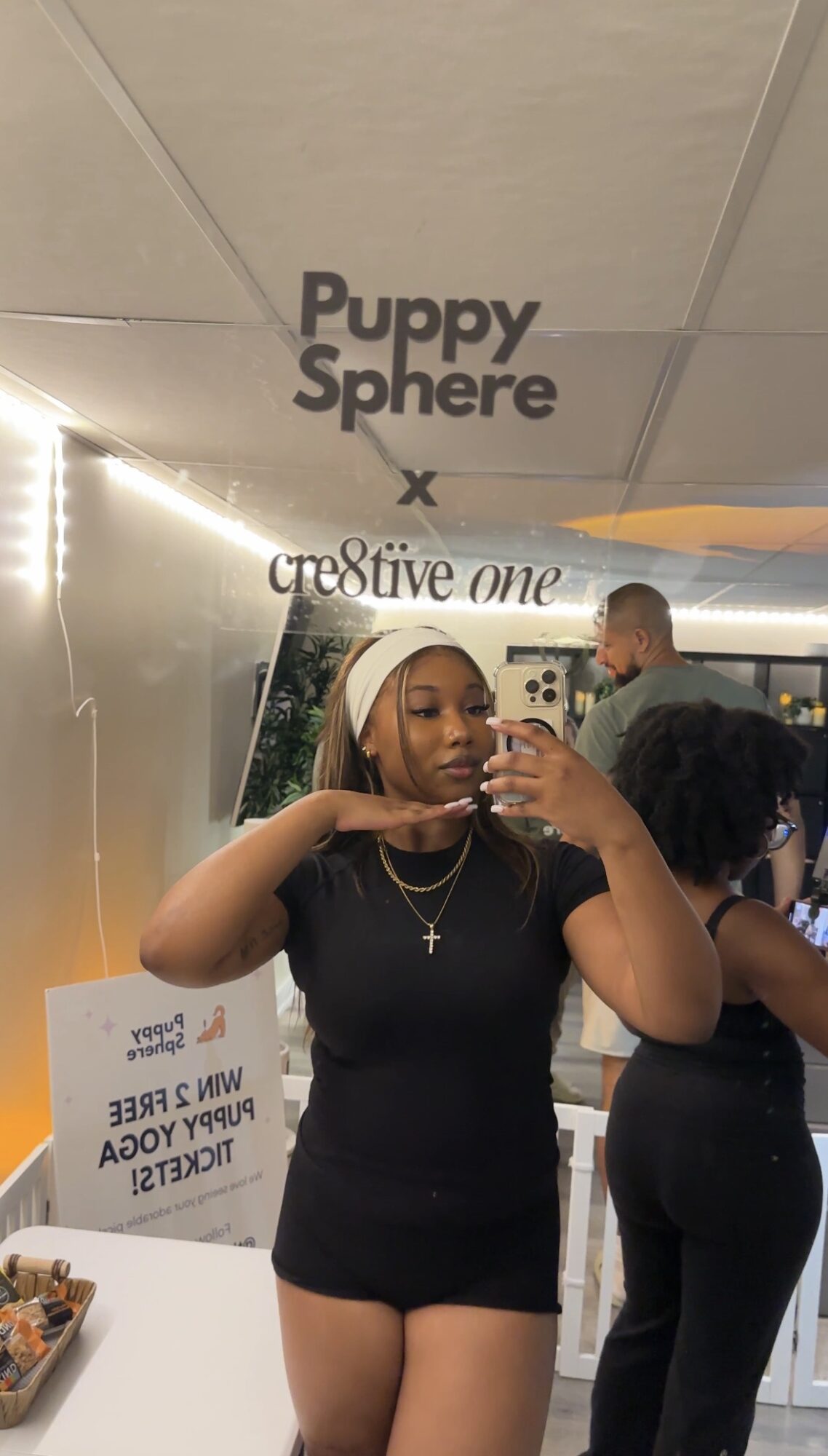 Young woman taking a selfie in a store, wearing a black outfit and a white headscarf, with other people in the background.