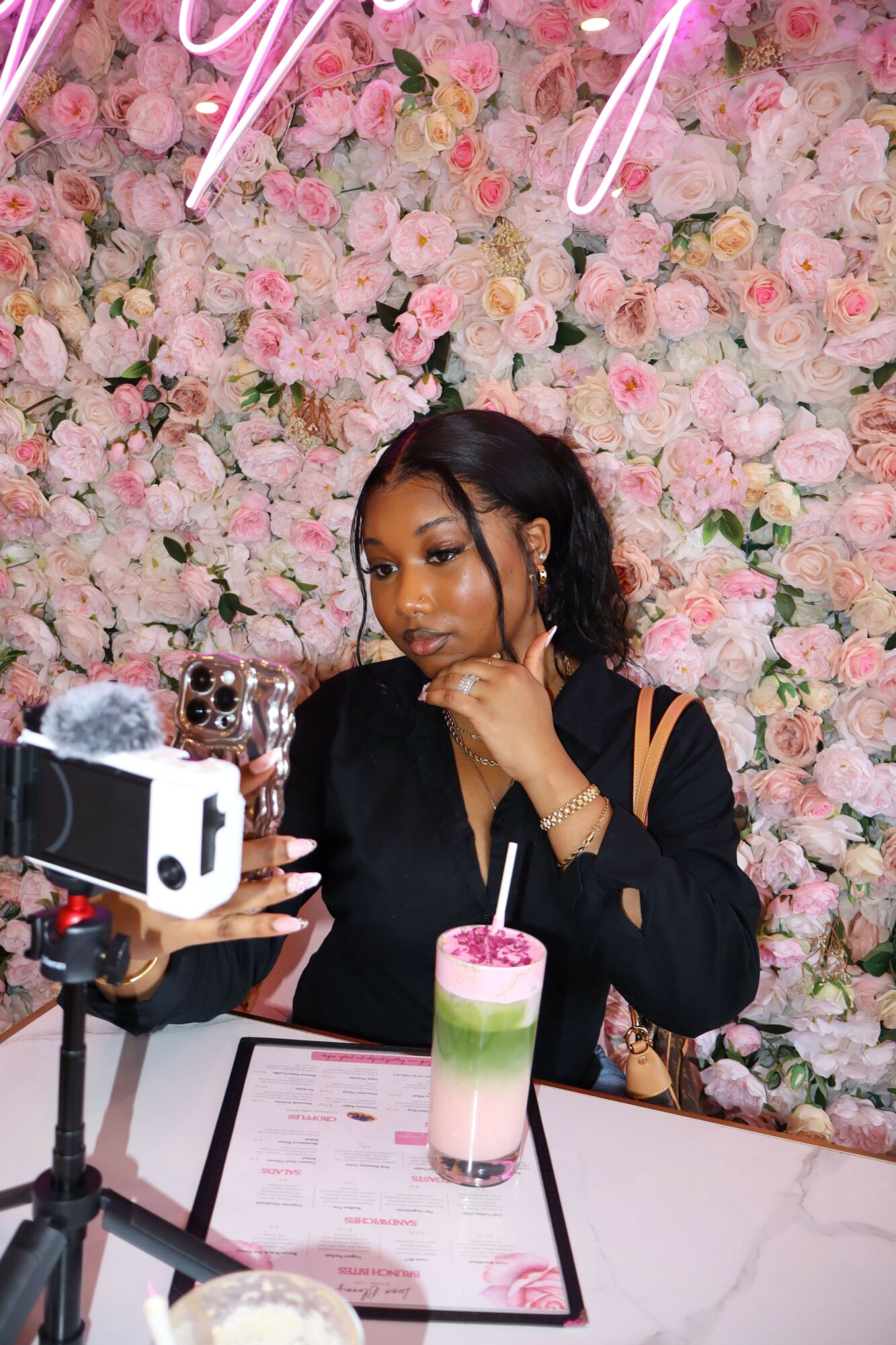 Woman sitting at a table with pink floral backdrop, drinking from a green and white cup, surrounded by cameras and documents.