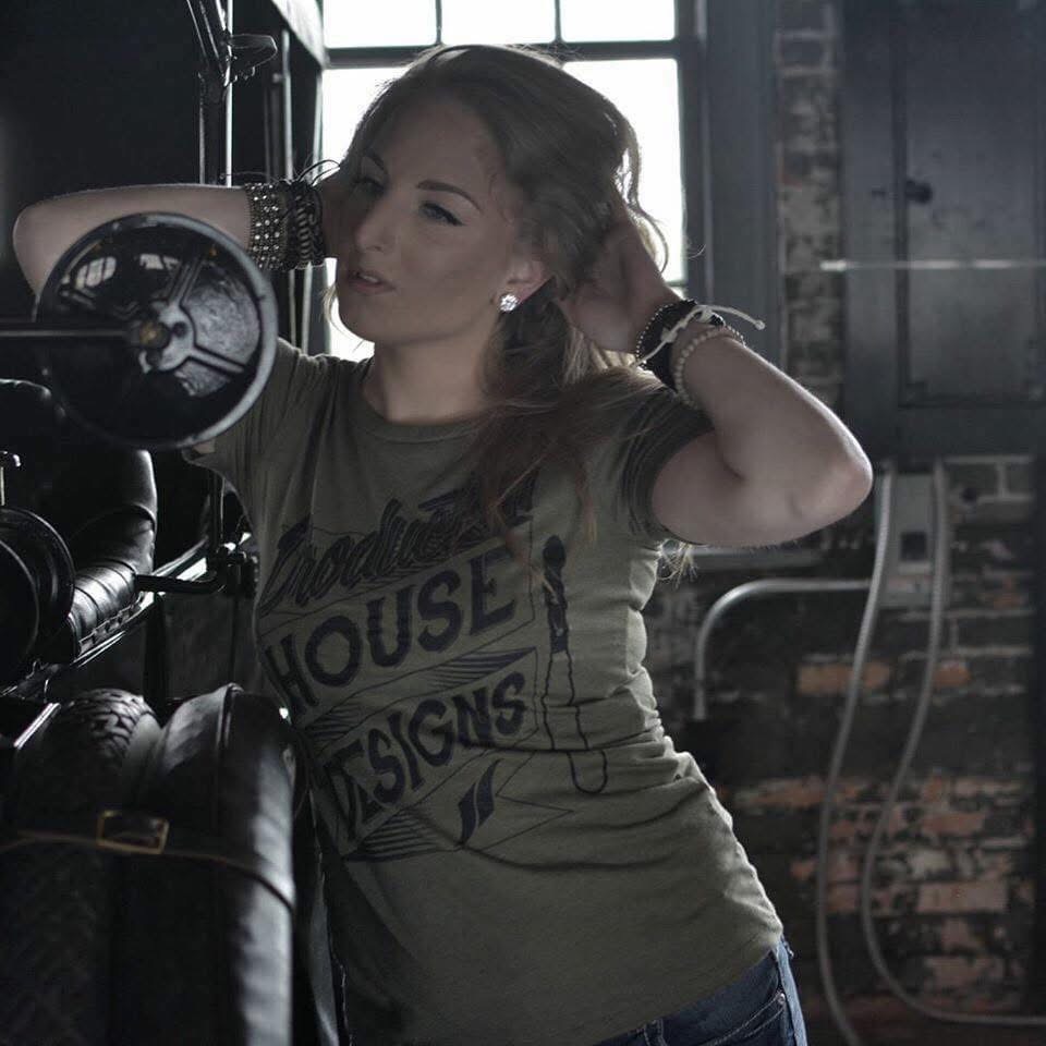 Woman with long hair adjusting her hair in a dimly lit industrial space, wearing a graphic t-shirt and jewelry.